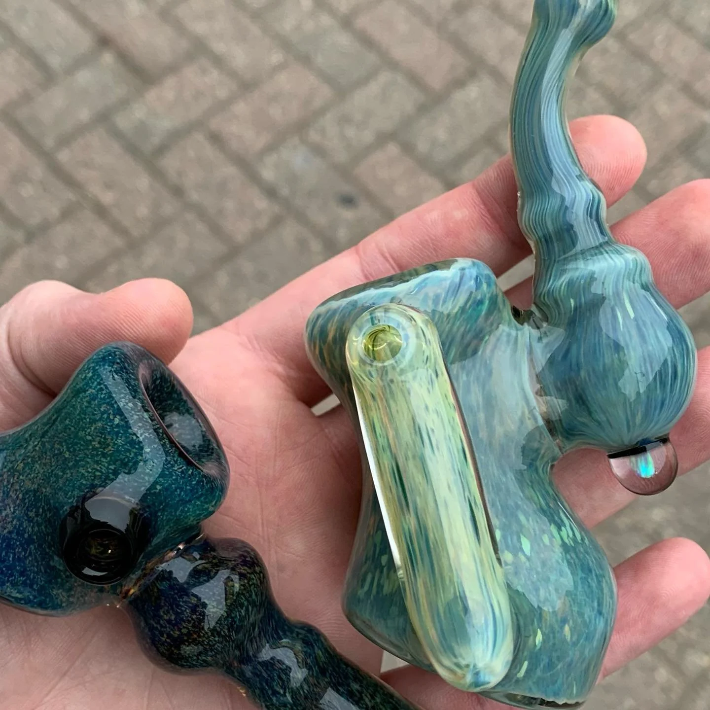 A person holding two colorful glass smoking pipes with a brick pavement in the background.