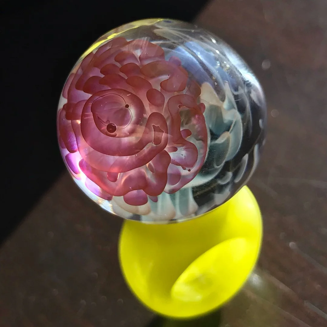 Close-up of a glass marble with a pink and purple internal swirl design, placed on a yellow stand.