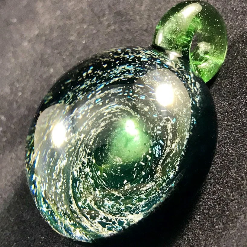 A glass paperweight with a galaxy design, featuring swirls of black, white, green, and blue, on a black surface.