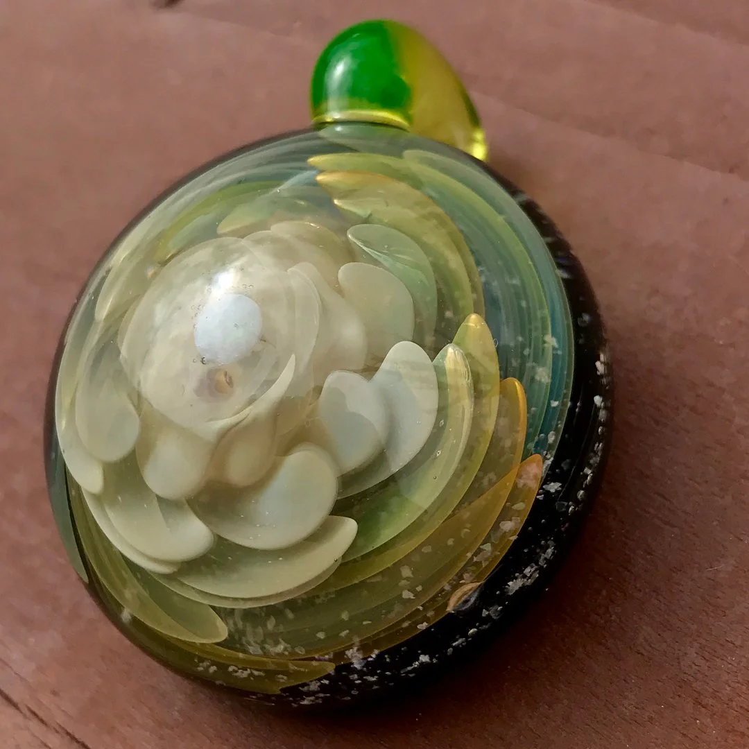 Close-up of a decorative glass paperweight resembling a blooming flower with layered petals in shades of white, yellow, and green, resting on a wooden surface.