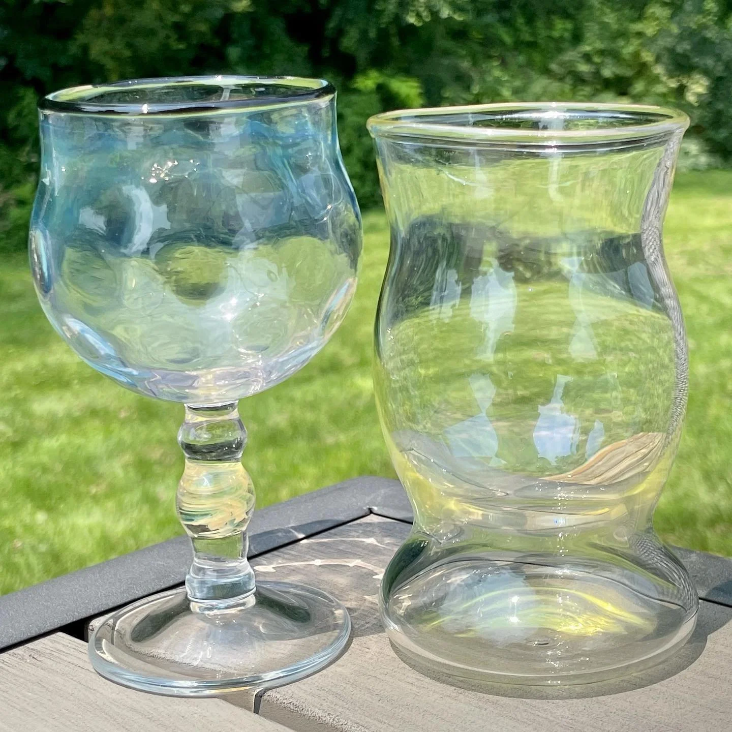 Two clear glass vases, one with a decorative stem, are placed outdoors on a wooden surface with a background of green grass and trees.