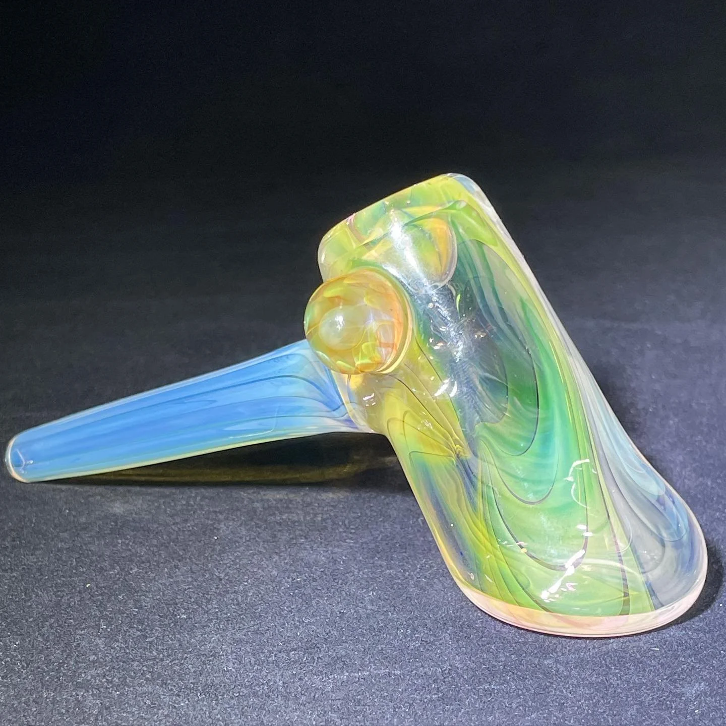Colorful glass art piece with a curved green and yellow body and a long blue tail set against a dark background.