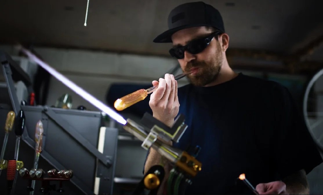 A man wearing didymium and a black cap is working in a glass blowing workshop, using a GTT Mirage torch to heat a glass object.