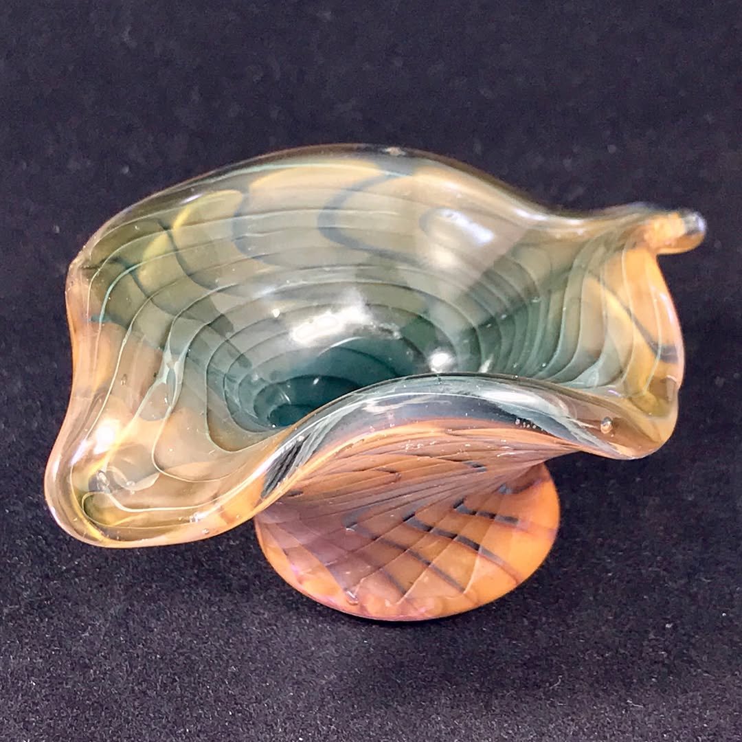 Colorful glass art piece with a green and gold swirl pattern, shaped like a bowl or vase, on a dark surface.