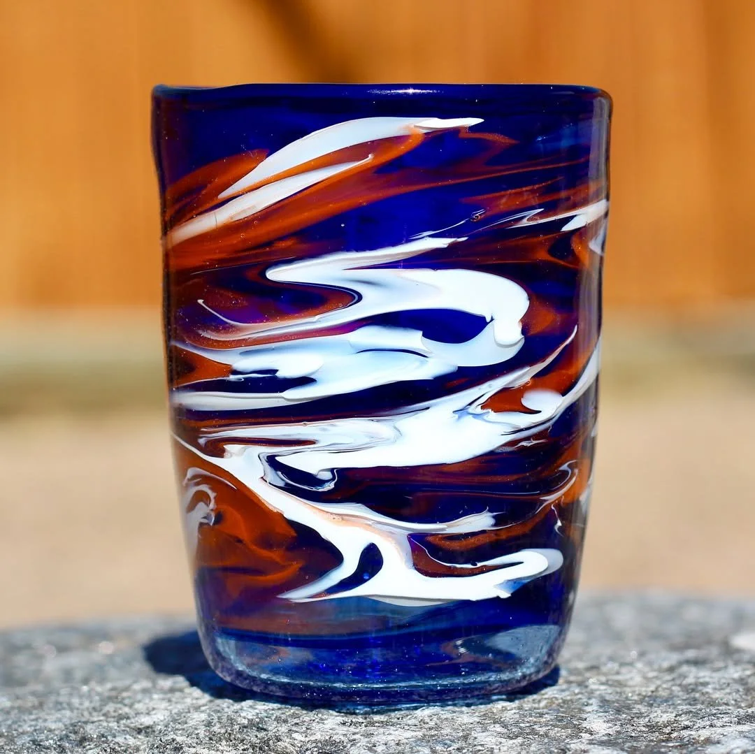 A colorful glass with blue, white, and brown swirled patterns, sitting on a rough stone surface with a blurred wooden background.