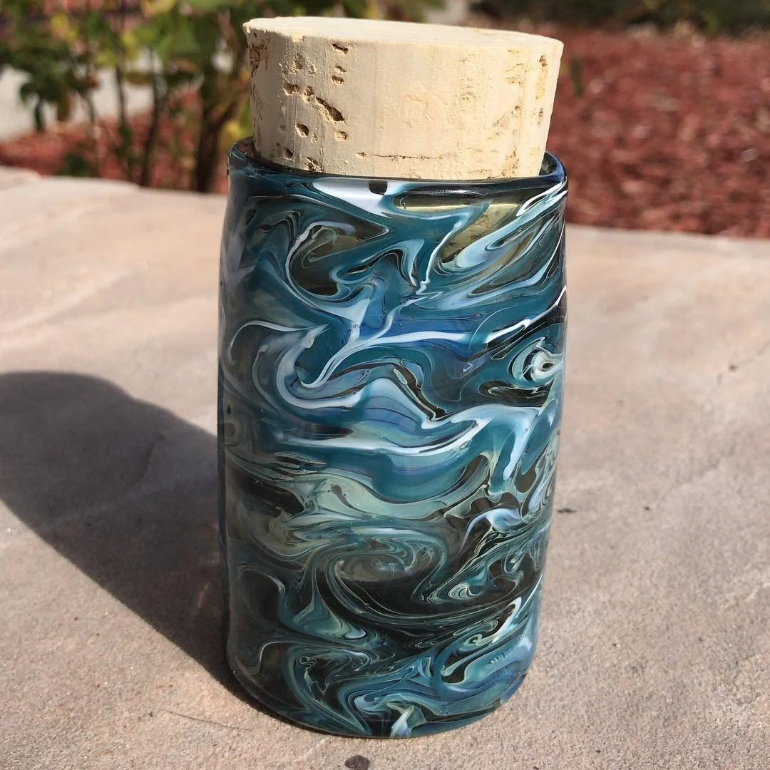 A glass container with a swirling blue, black, and white pattern, topped with a cork stopper, placed on a concrete surface outdoors.