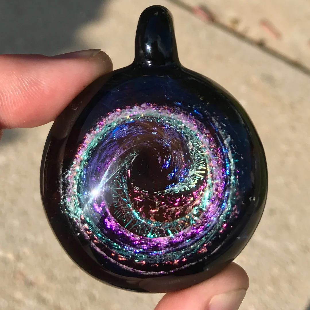 A hand holding a black pendant with a swirling galaxy design containing colorful, glittering lights and patterns.