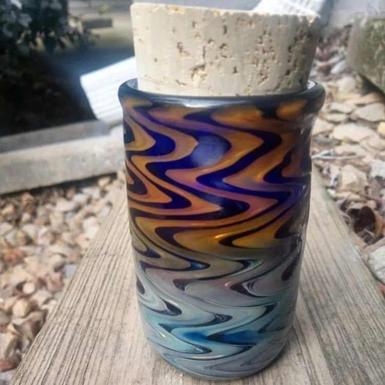 A colorful glass vase with a swirling design holding a potted plant, placed on a wooden surface outside.
