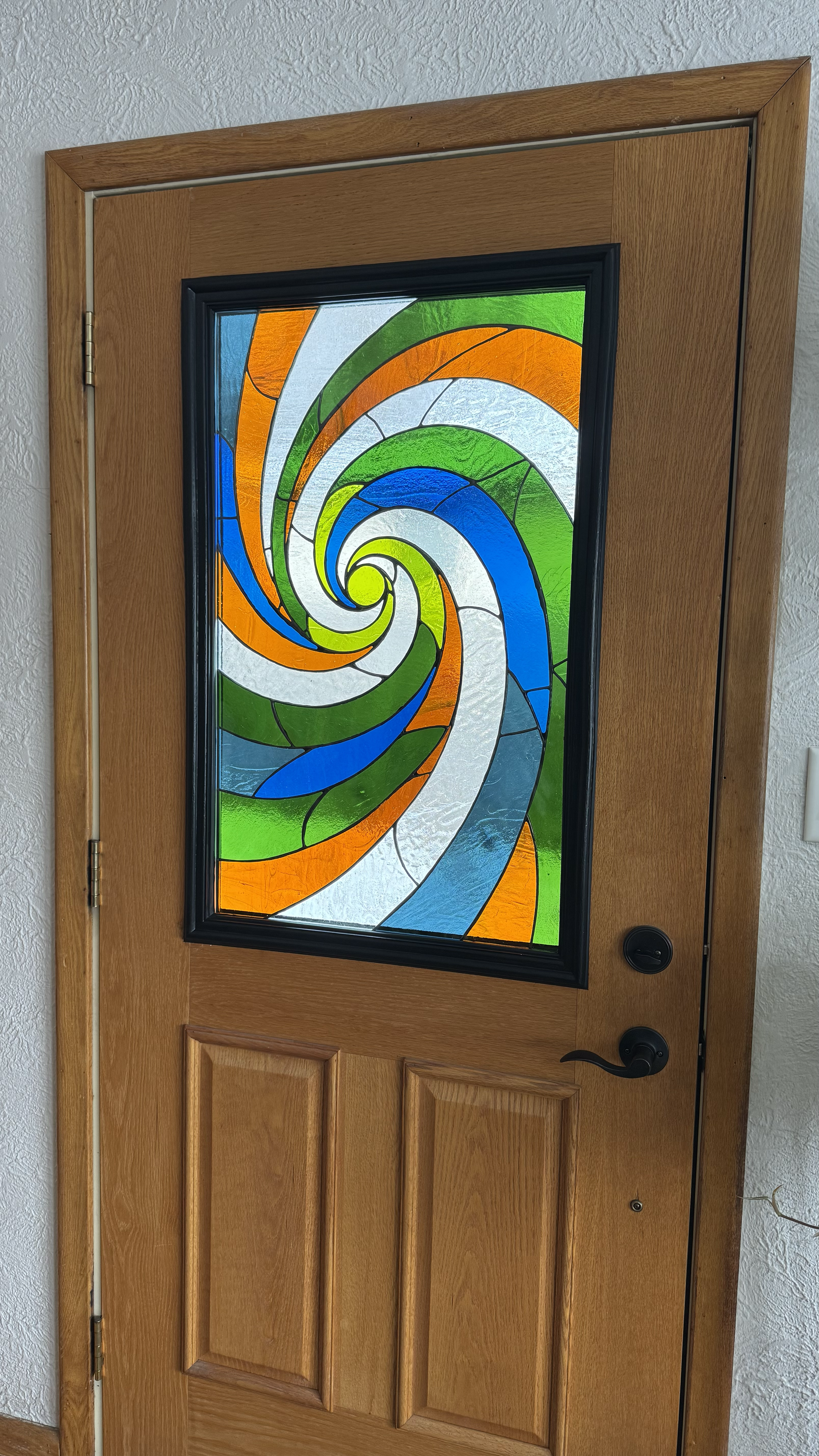 Wooden front door with a colorful stained glass window featuring a swirling pattern in green, blue, orange, yellow, and white.