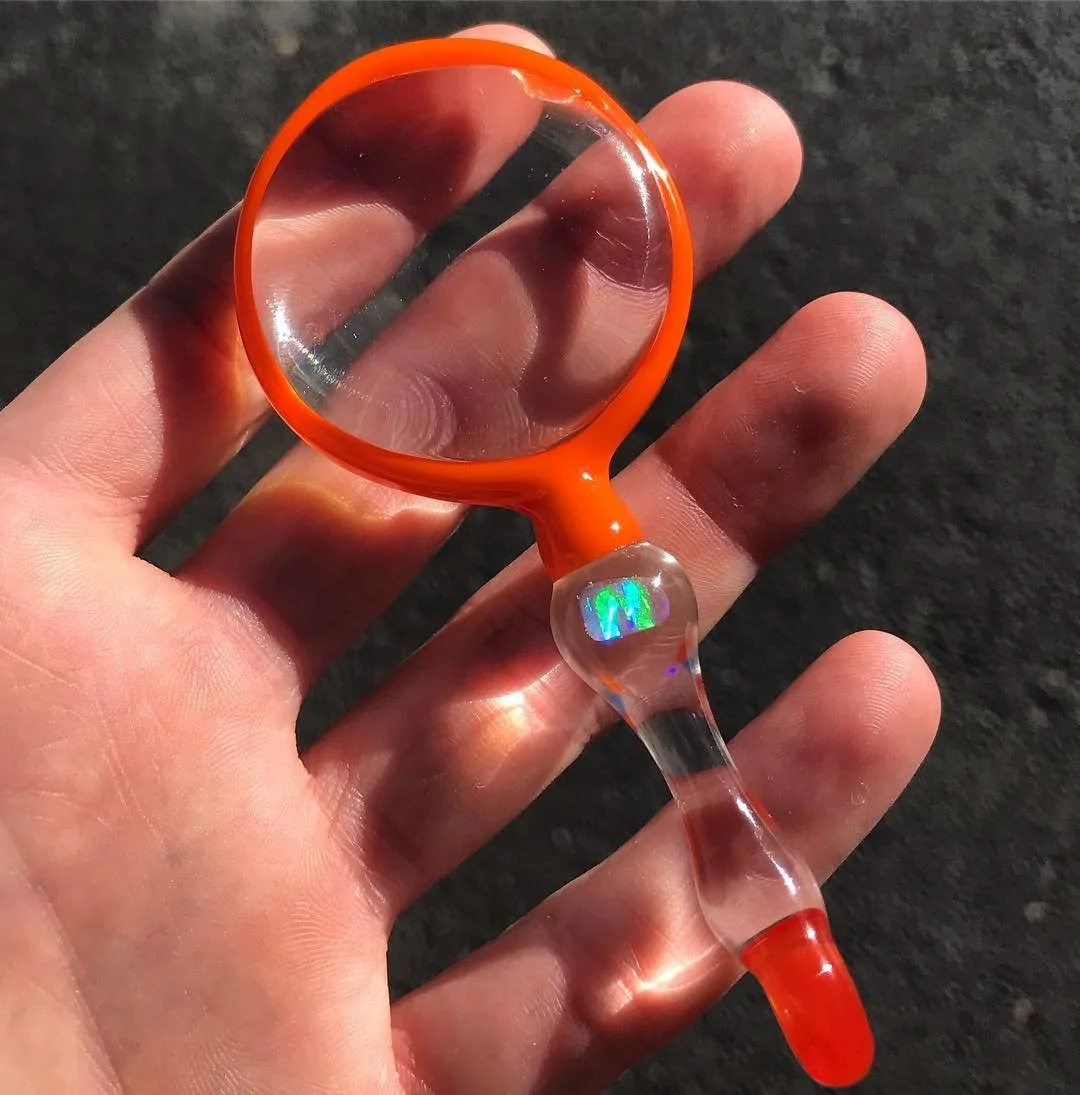 A small magnifying glass with an orange rim and handle, held in a person's hand, with a rainbow-colored holographic sticker on the handle, against a dark background.