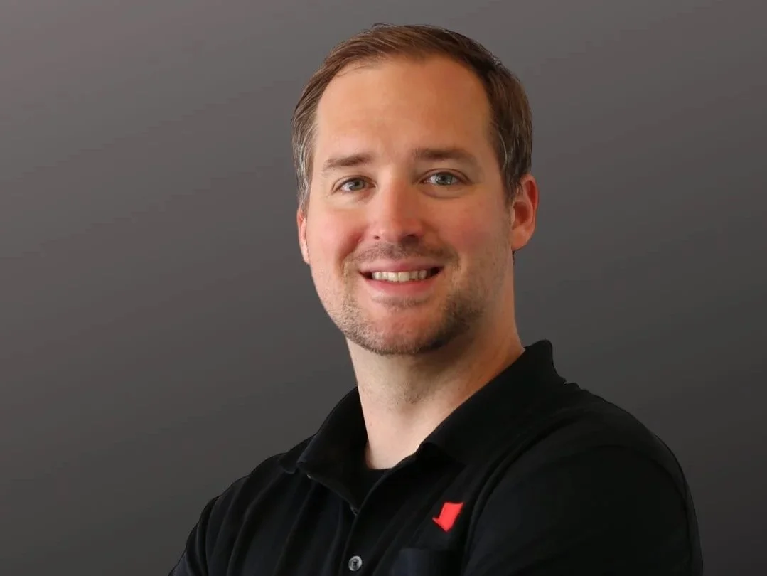 A smiling man with short brown hair, light skin, and a goatee, wearing a black collared shirt, standing against a gray background.