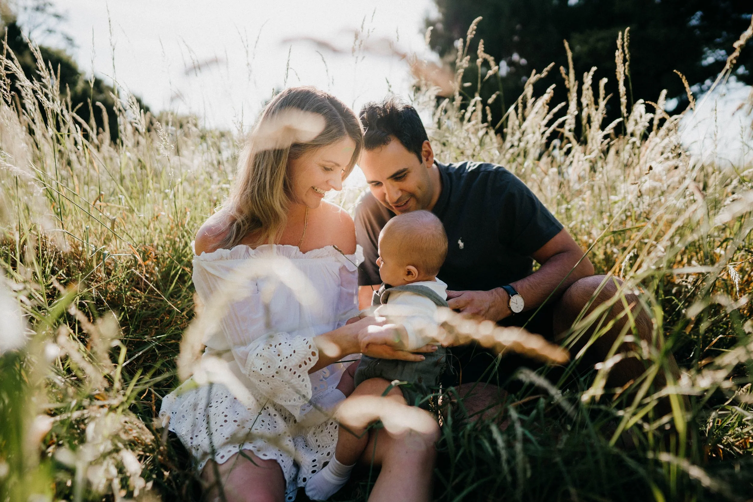 A couple sitting in a grassy field holding a baby, enjoying a sunny day.