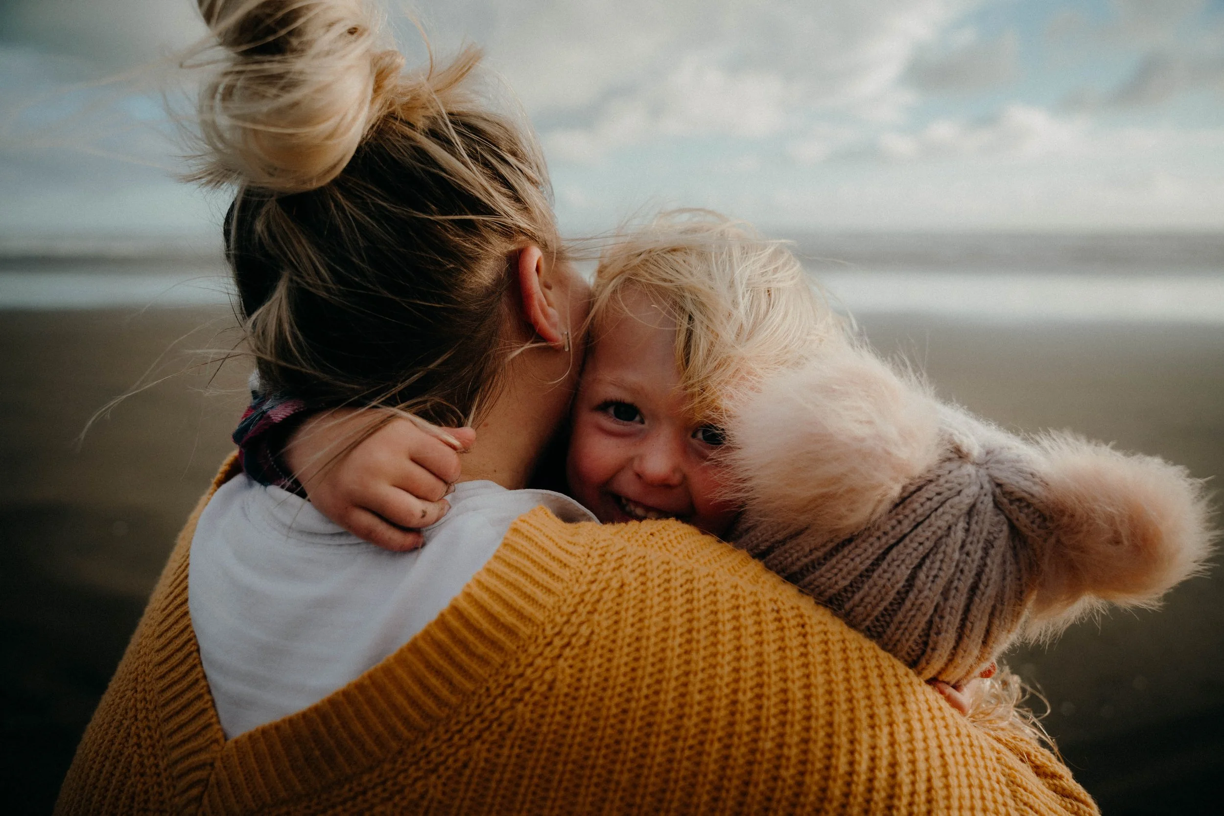 A woman with a bun hairstyle hugs a smiling child wearing a warm knit hat with pom-poms, on a beach.