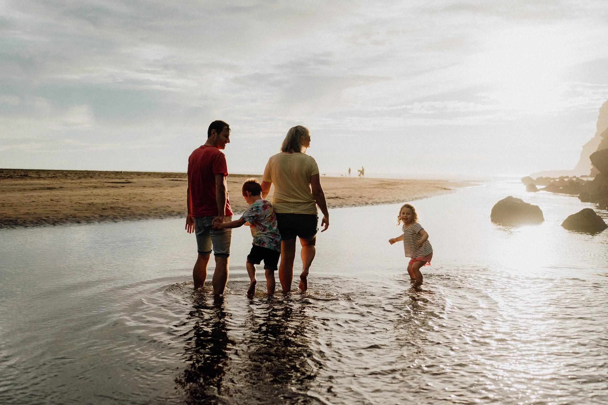 Family of four wading in shallow water on a beach during sunset.