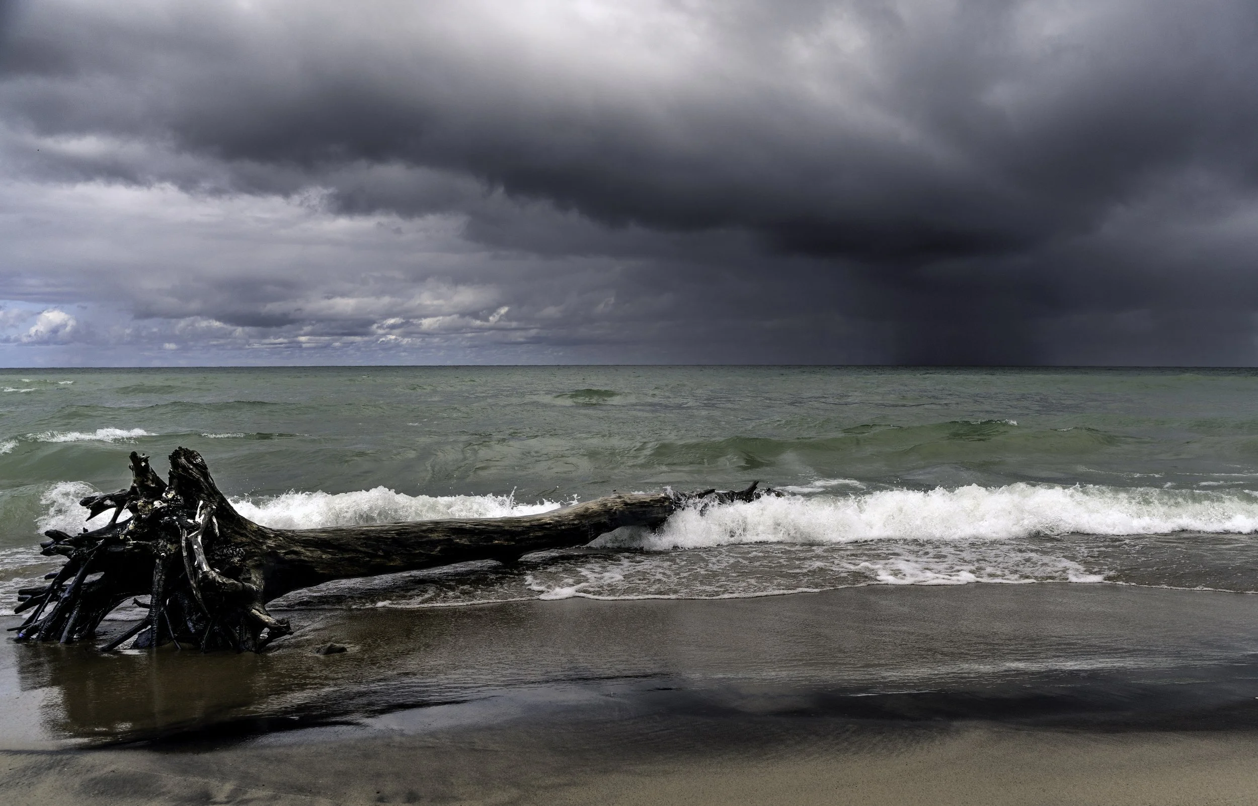 Stormy sky over the ocean with dark clouds, turbulent waves, and a fallen driftwood log on the sandy beach.