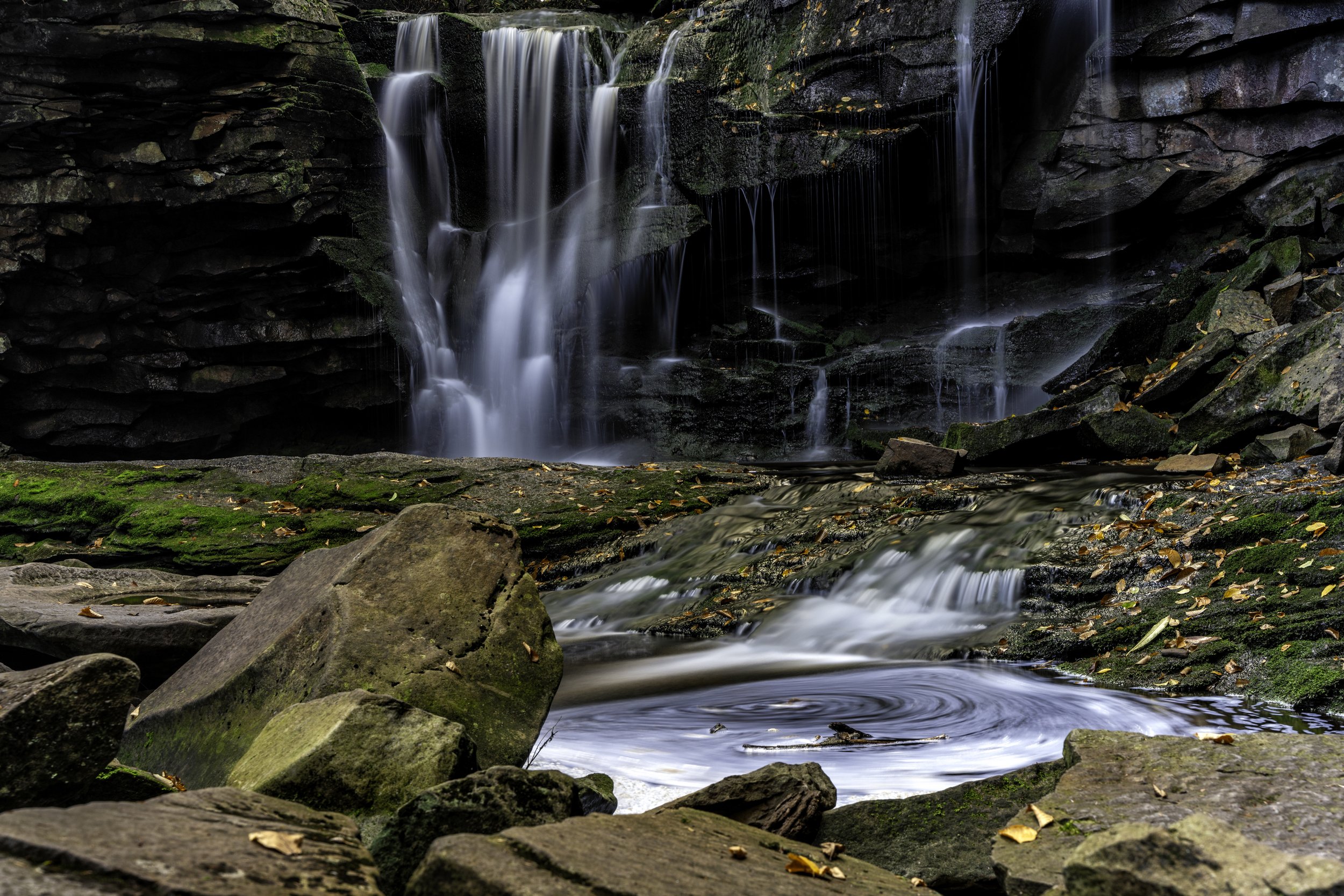 A small waterfall flowing over rocks into a stream surrounded by moss-covered stones and fallen leaves.