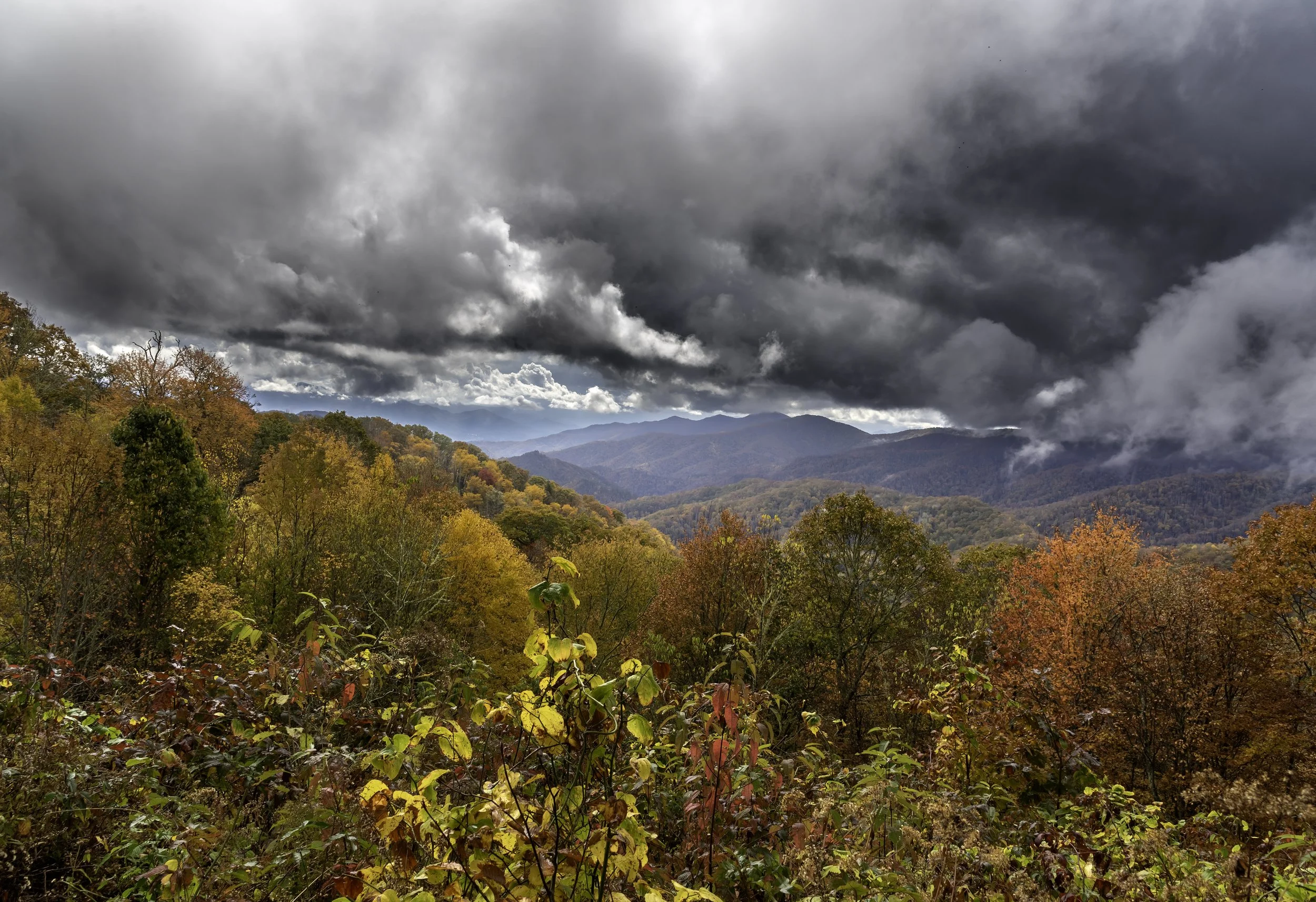 A scenic view of a forested mountain landscape during autumn with colorful fall foliage, under a stormy sky with dark, heavy clouds.