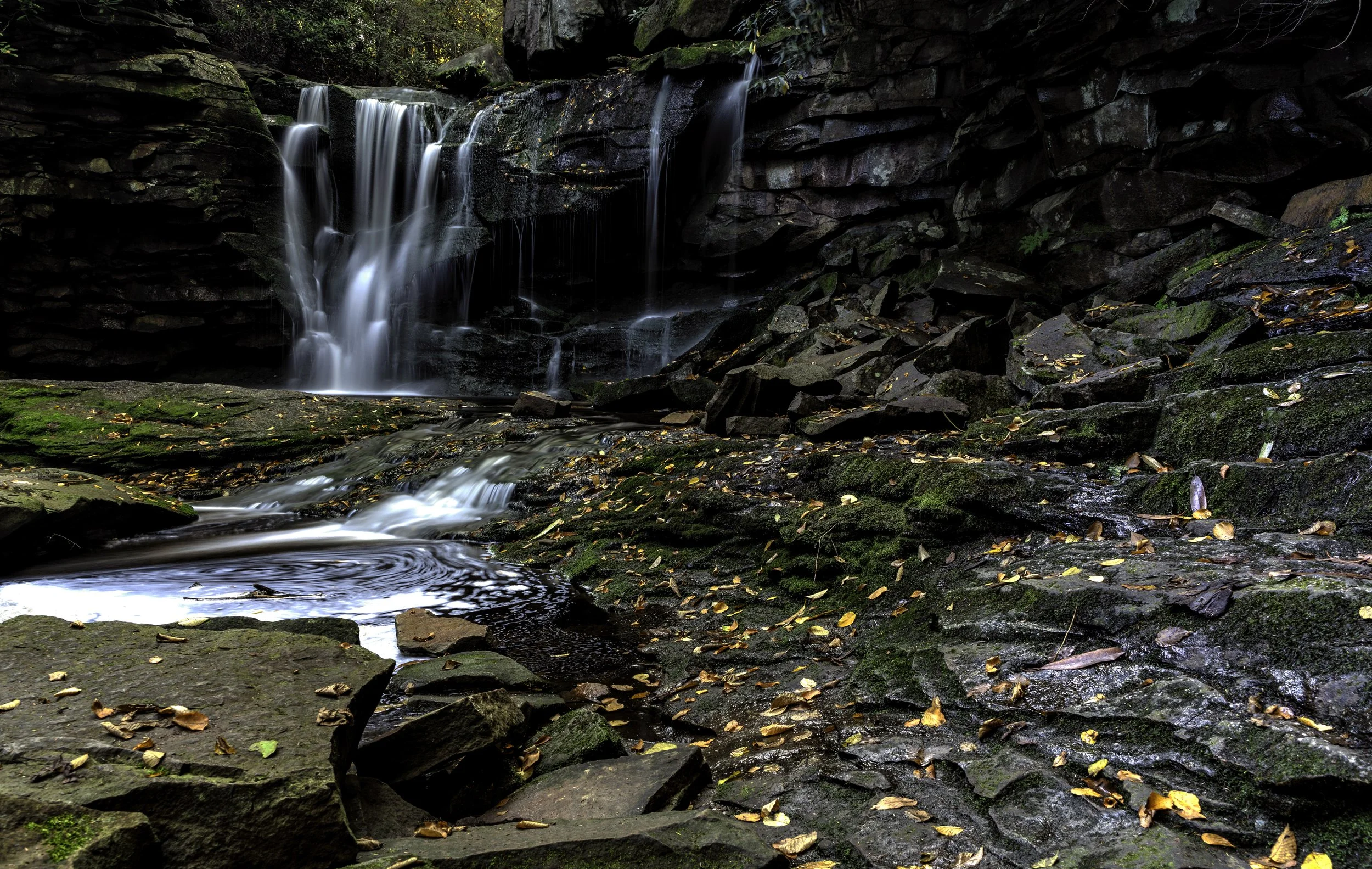 A small waterfall cascading over rocks in a forested area with moss and fallen leaves