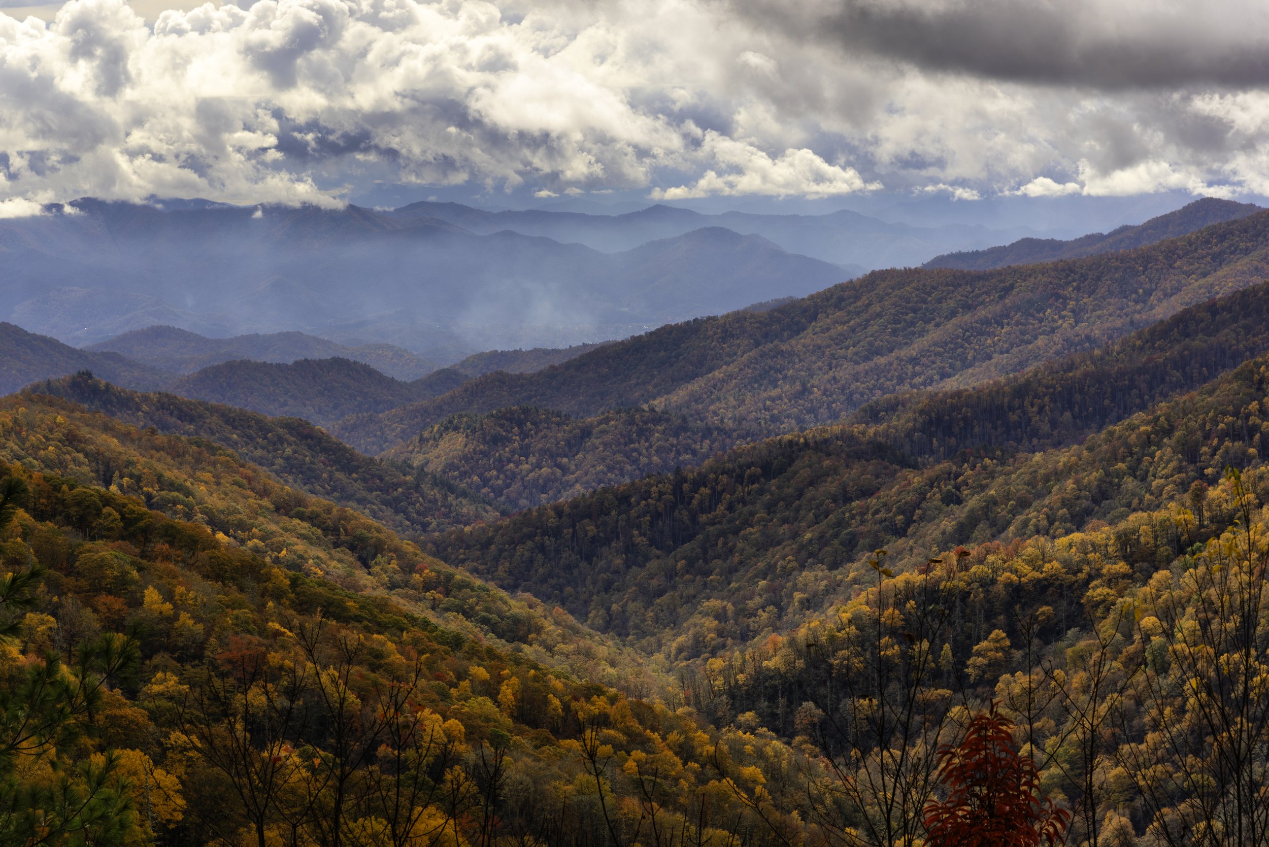 A view of mountains covered in autumn foliage with trees in shades of orange, yellow, and red, under a cloudy sky with patches of blue.