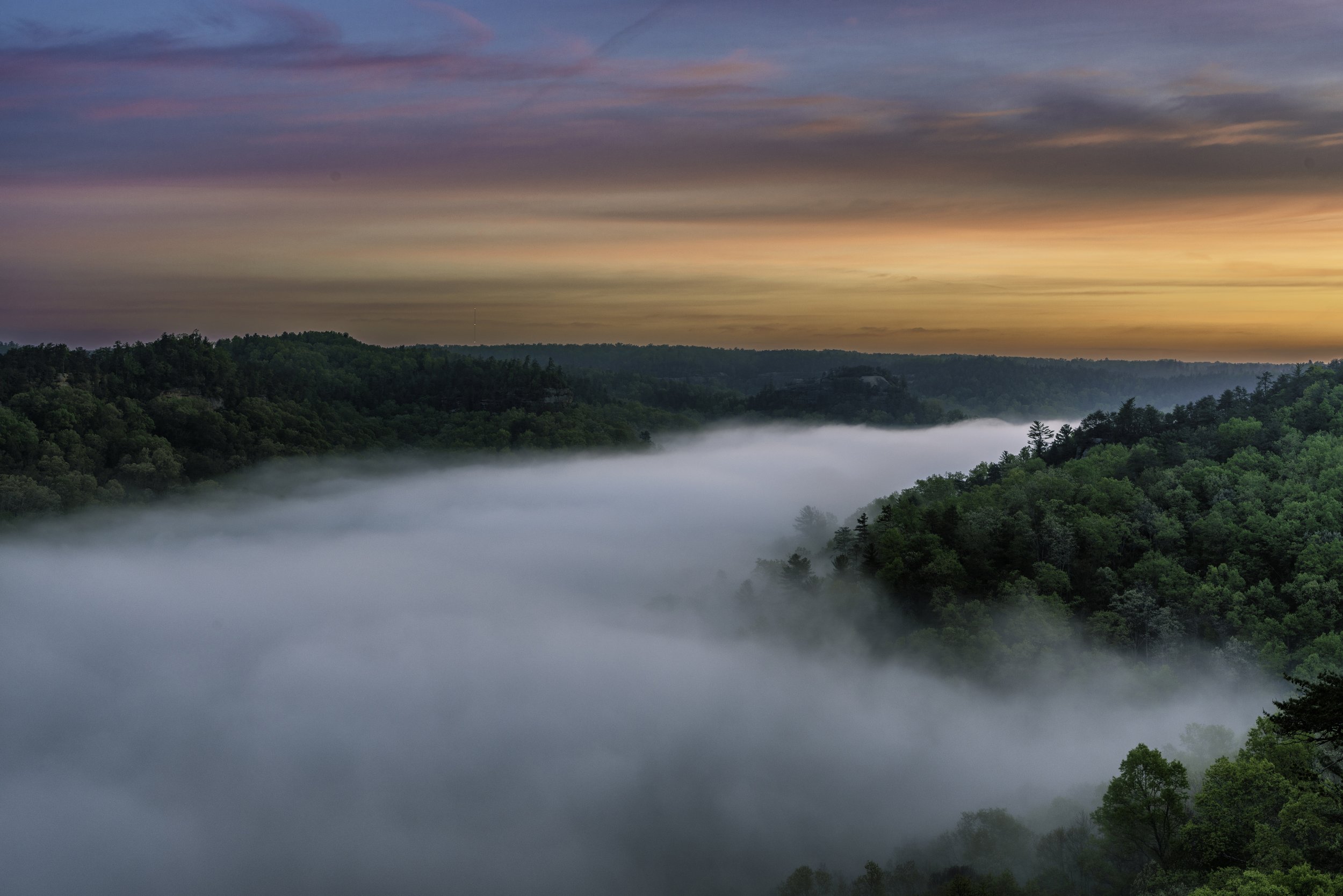 Sunrise over a mountainous landscape with a layer of fog covering the valleys and a colorful sky with clouds.