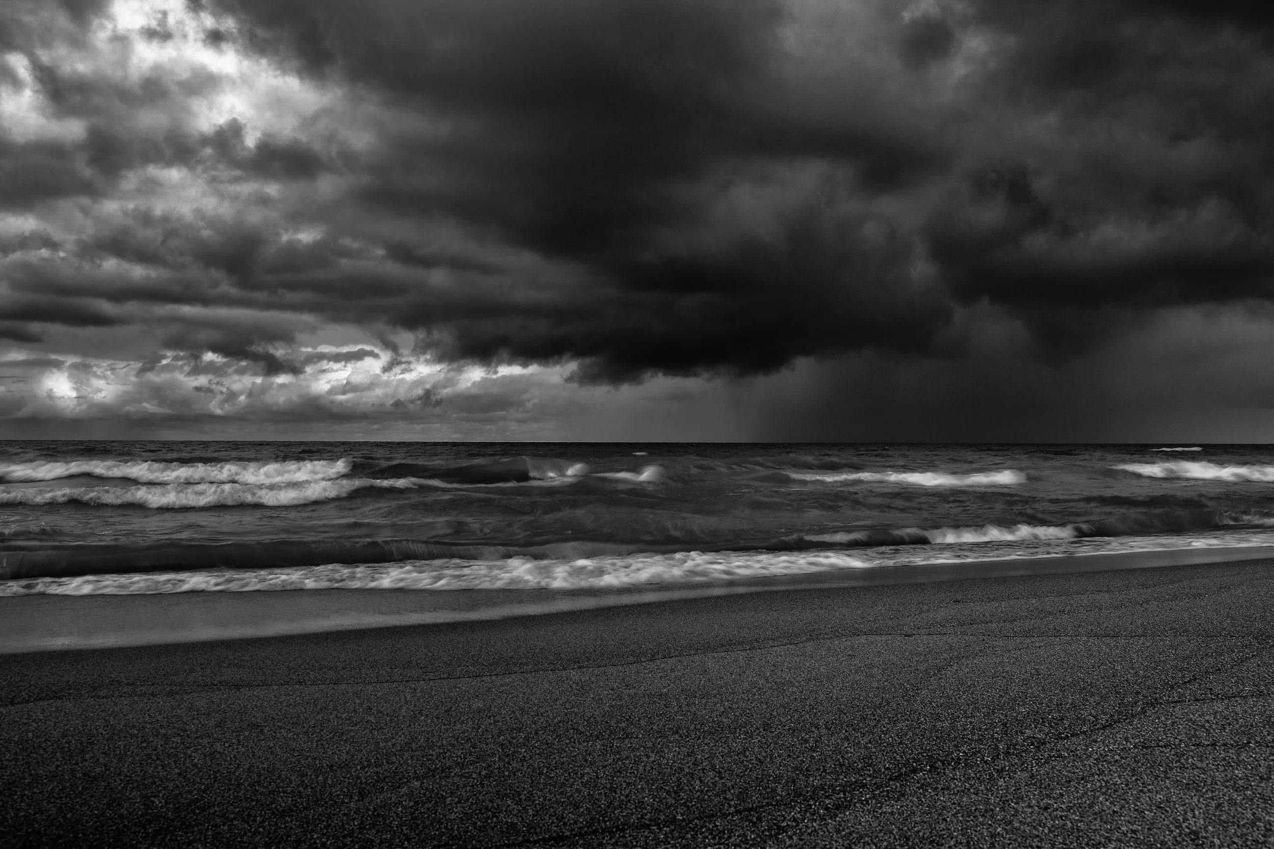 Black and white photo of dark storm clouds over an ocean with waves crashing onto a sandy beach.