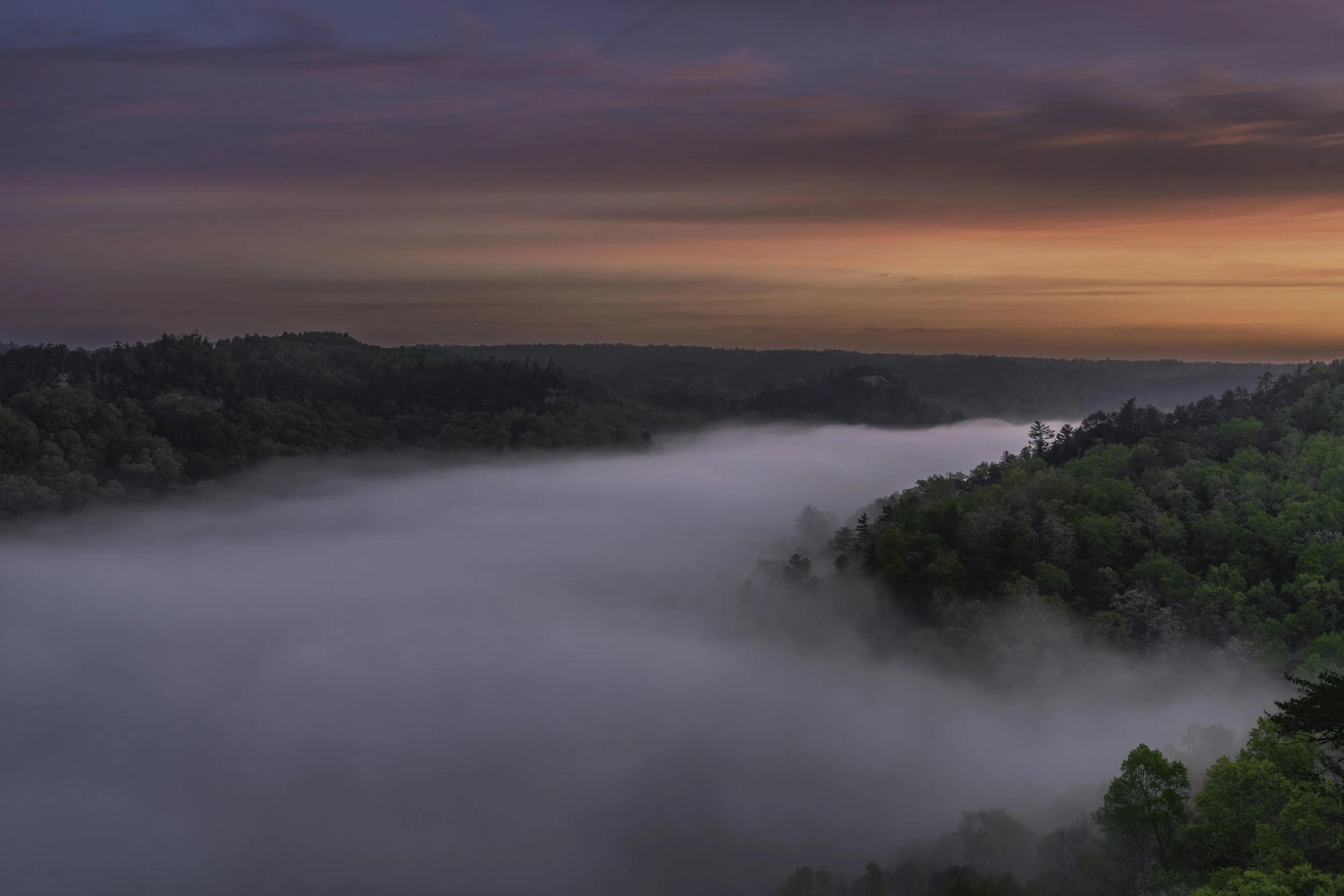 A scenic landscape of a valley filled with fog, surrounded by green hills and trees, with a colorful sunset sky in the background.