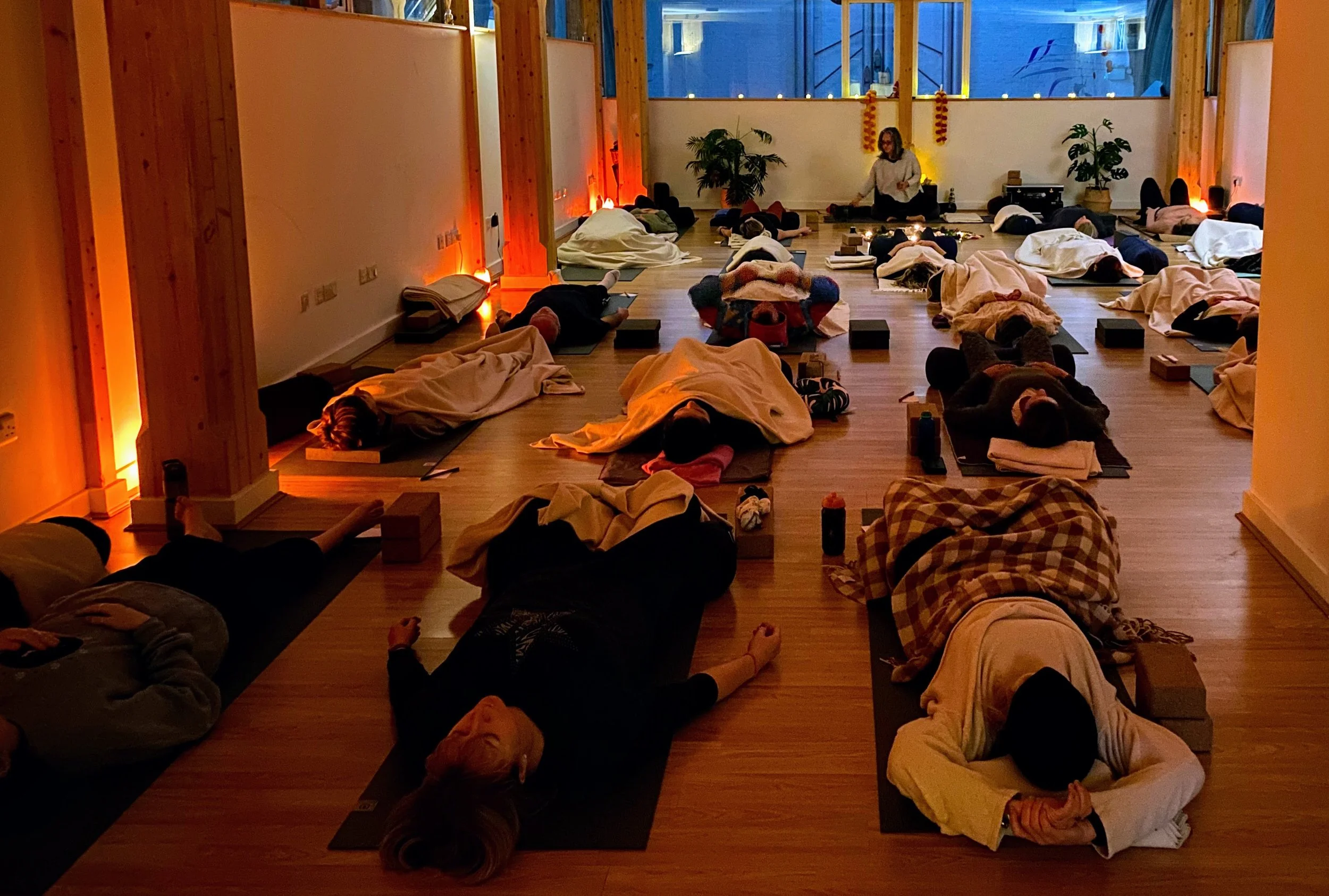 A group of people lying on yoga mats in a dimly lit room participating in a yoga or meditation class, with a instructor at the front of the room.