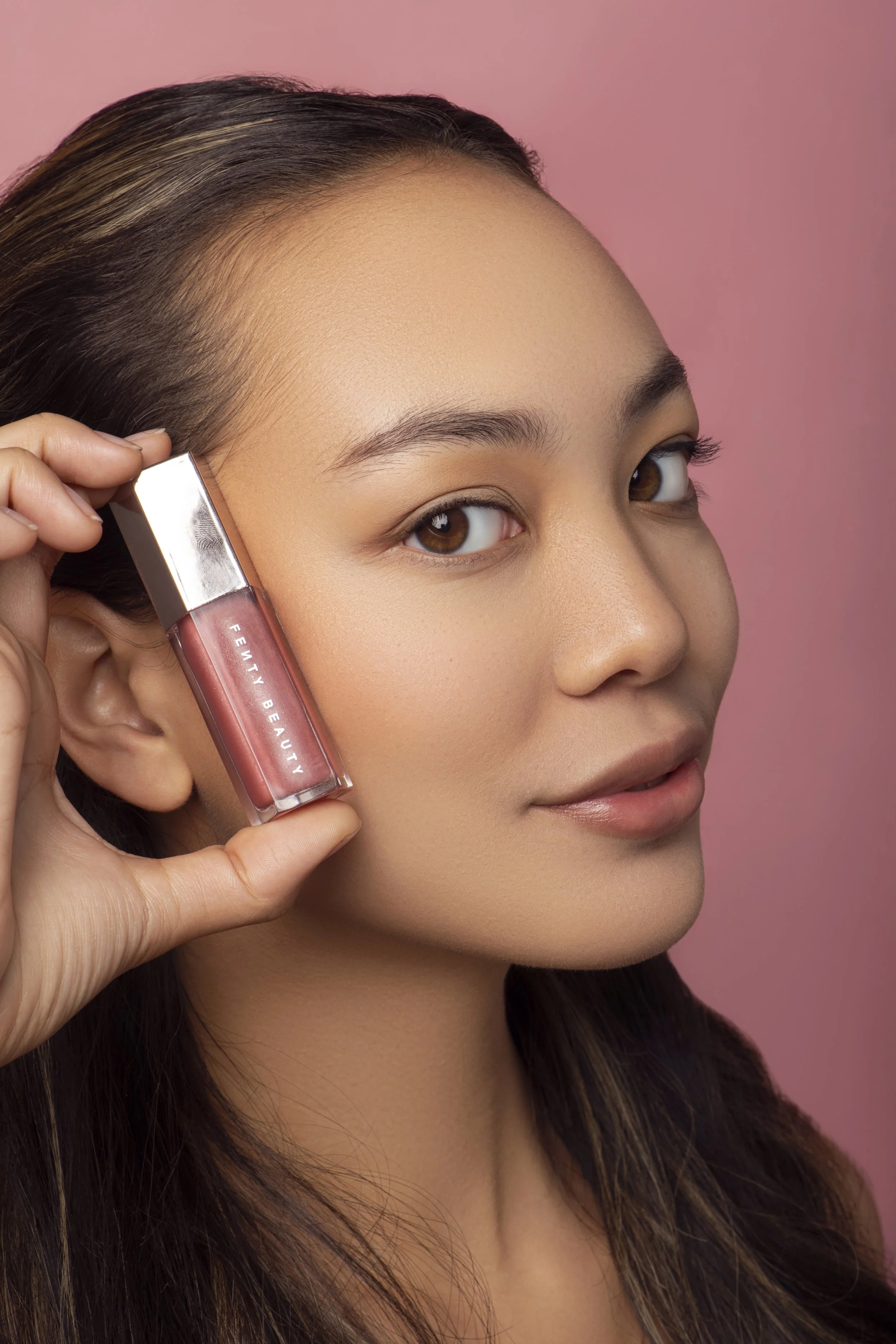 Close-up of a woman with long dark hair holding a gloss lipstick near her cheek against a pink background.
