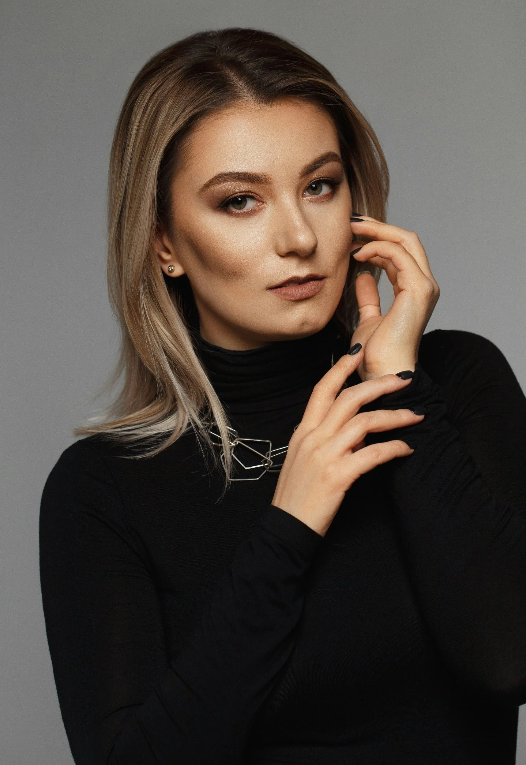 A woman with shoulder-length blonde hair, wearing a black turtleneck and silver geometric necklace, poses with her hands near her face against a neutral gray background.