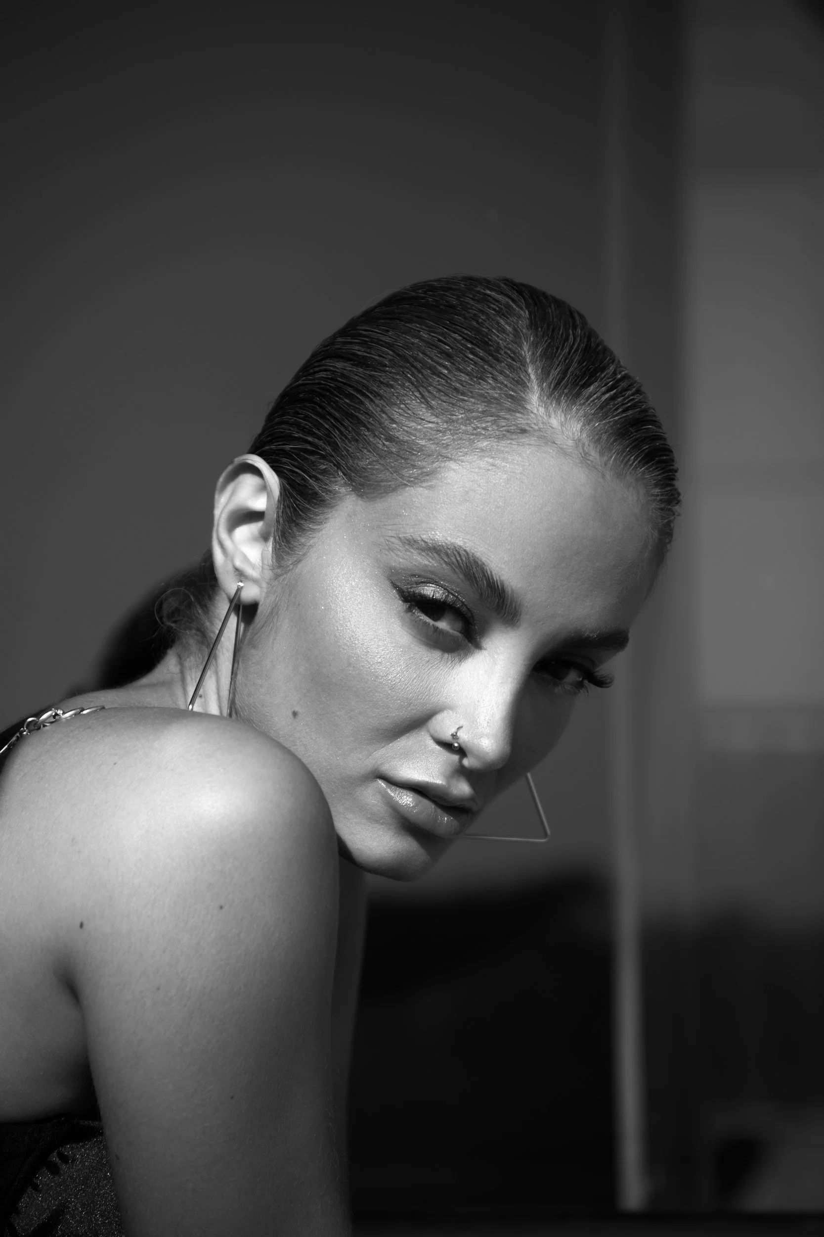 Black and white photo of a woman with slicked back hair, large hoop earrings, and a nose piercing, looking over her shoulder.