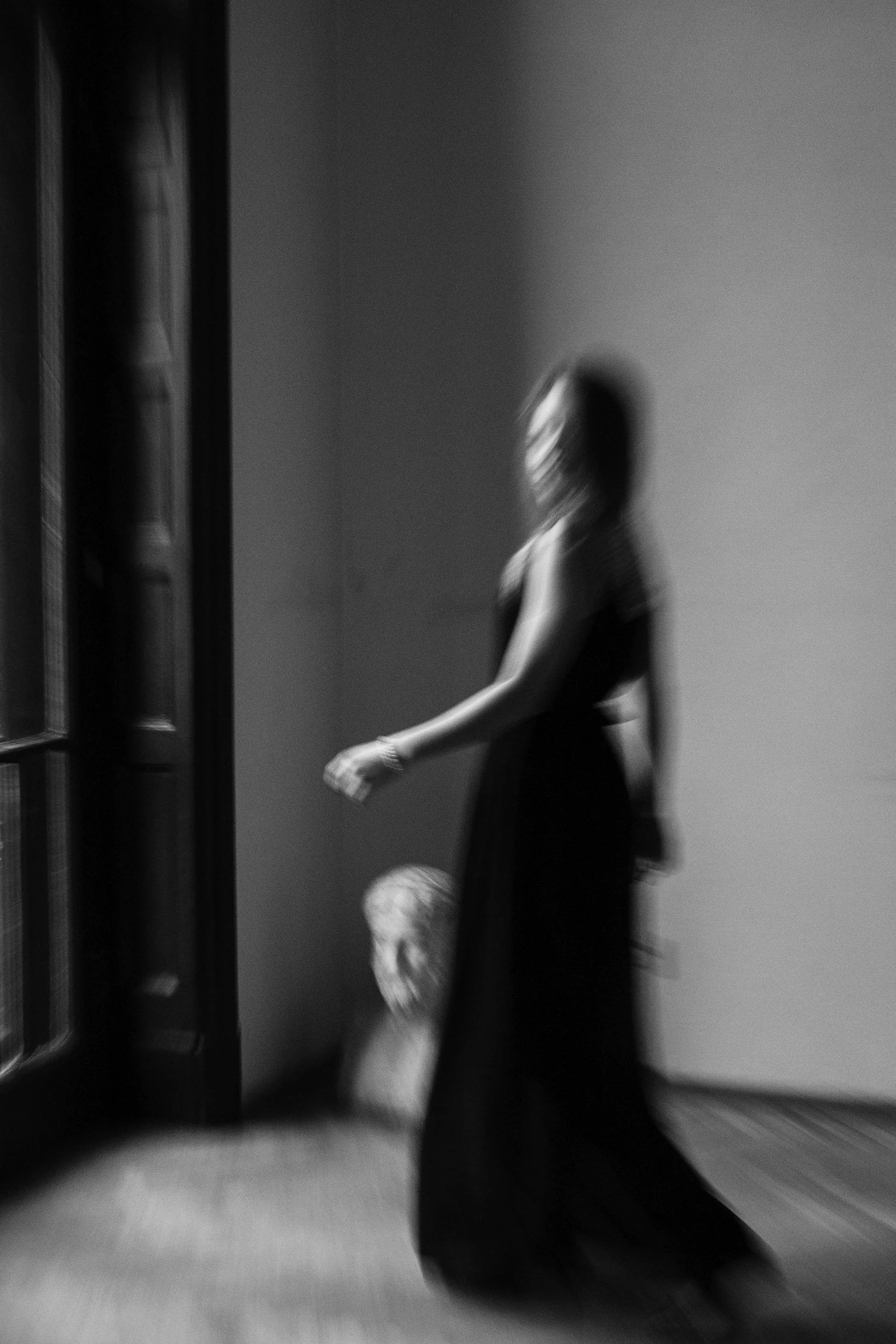 Black and white photo of a blurred woman in a long dress standing near a window, with a ceramic bust on the floor beside her.