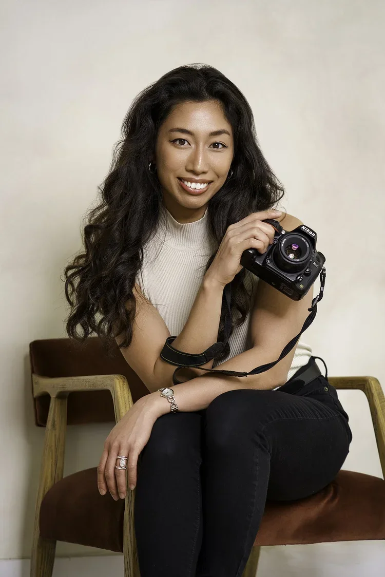 A young woman with long, curly dark hair smiling and holding a camera while sitting on a wooden and fabric chair against a plain wall.