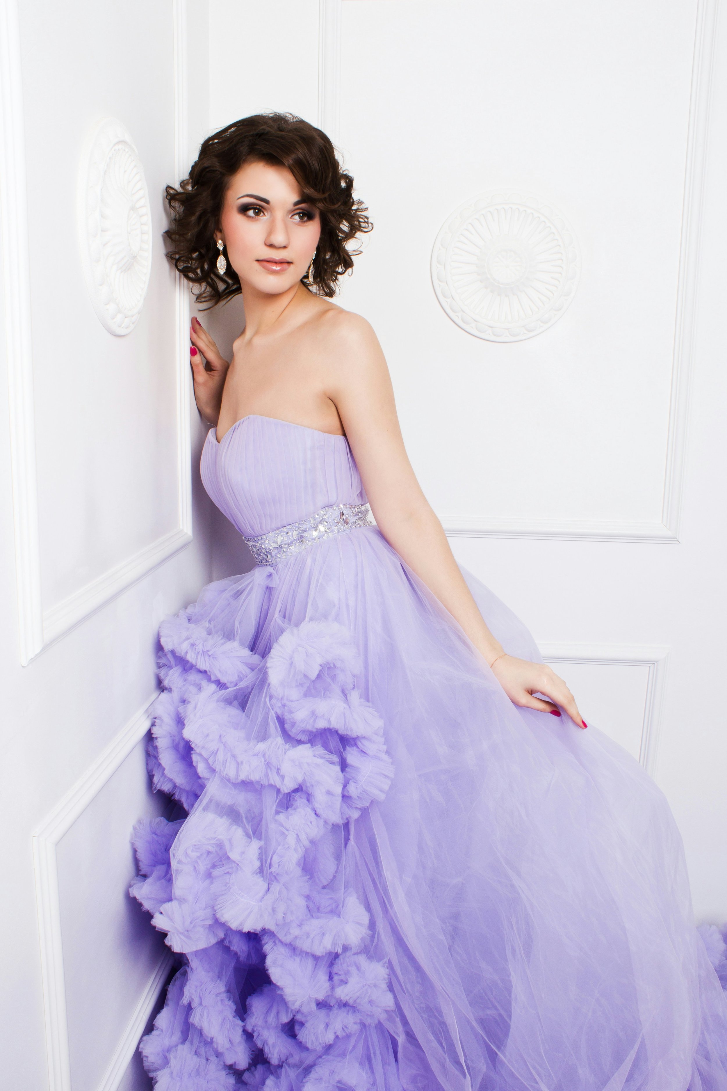 A woman wearing a strapless lavender gown with ruffled and tulle details, posing against a white wall with decorative molding.