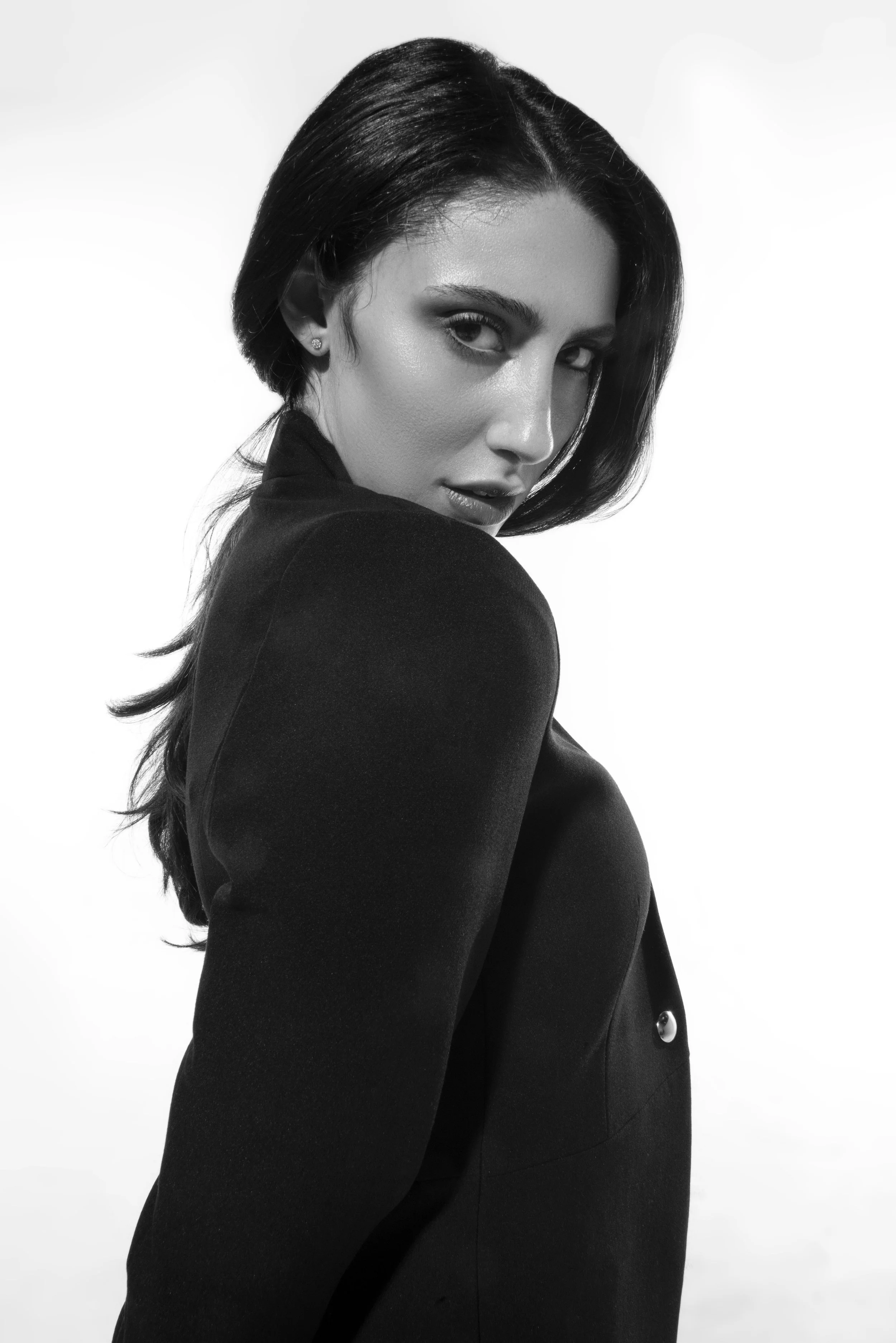 Black and white portrait of a woman with short dark hair, wearing a dark blazer, looking over her shoulder at the camera with a confident expression.