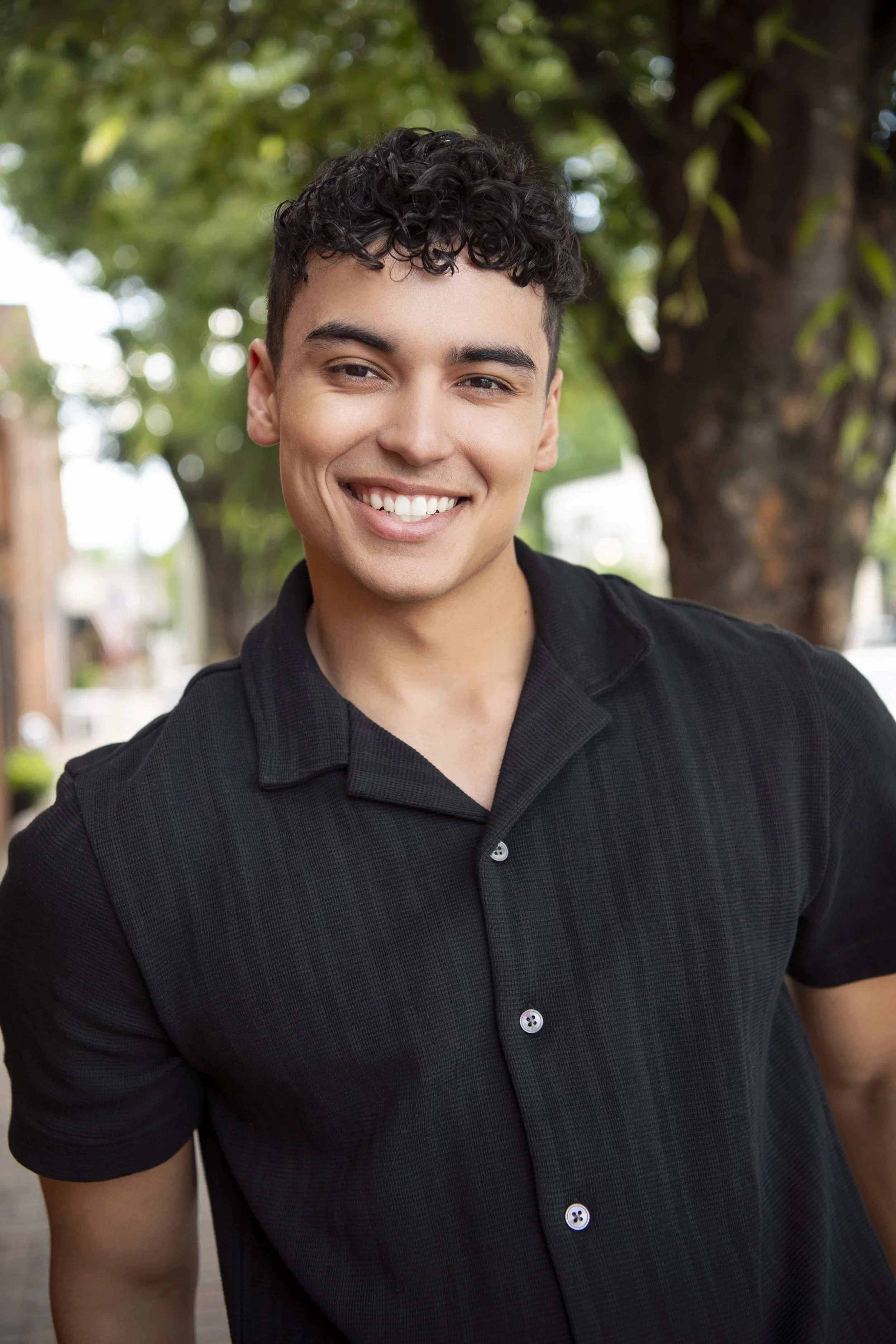 Portrait of a young man with curly black hair, smiling, wearing a black Button-up shirt, standing outdoors with a background of trees and blurred greenery.