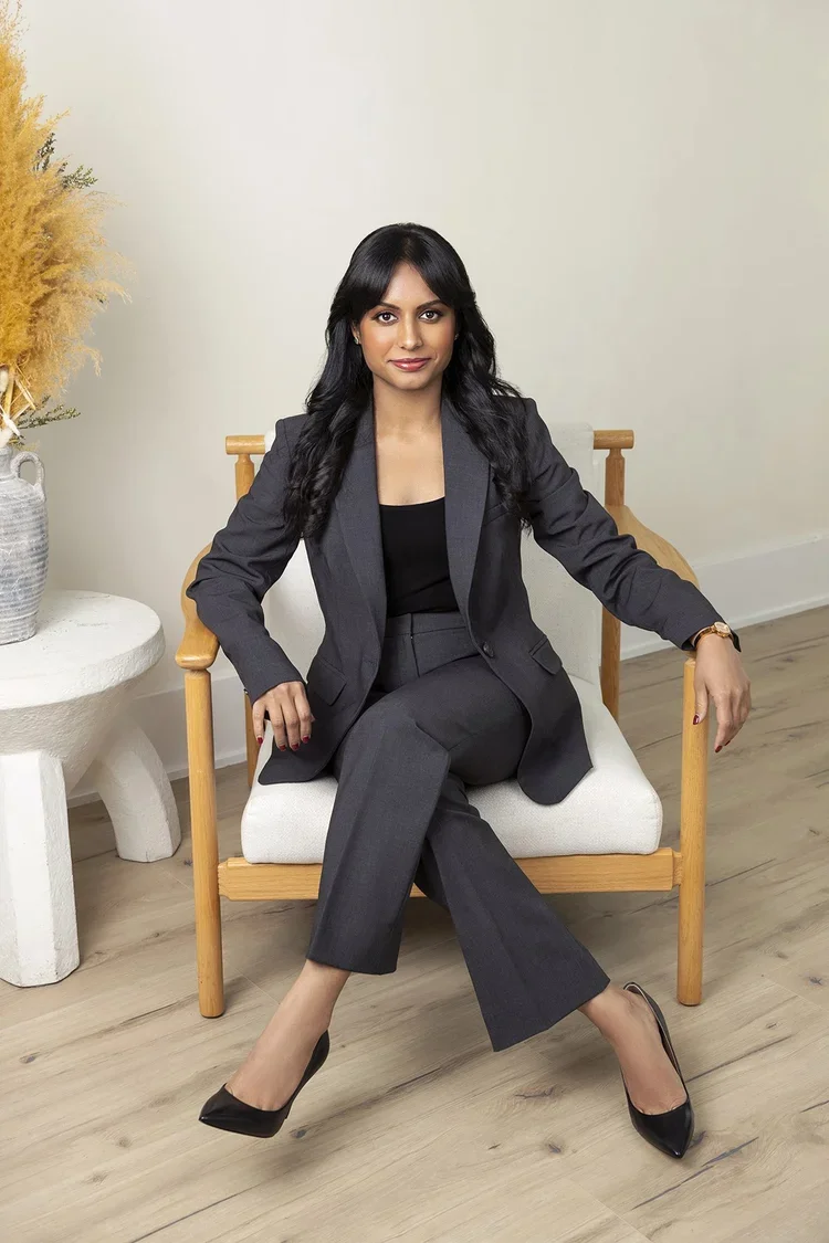 A woman in a dark gray business suit sitting on a light-colored chair in a room with wooden flooring and light-colored walls. There is a white side table with a decorative vase and dried plants to her left.