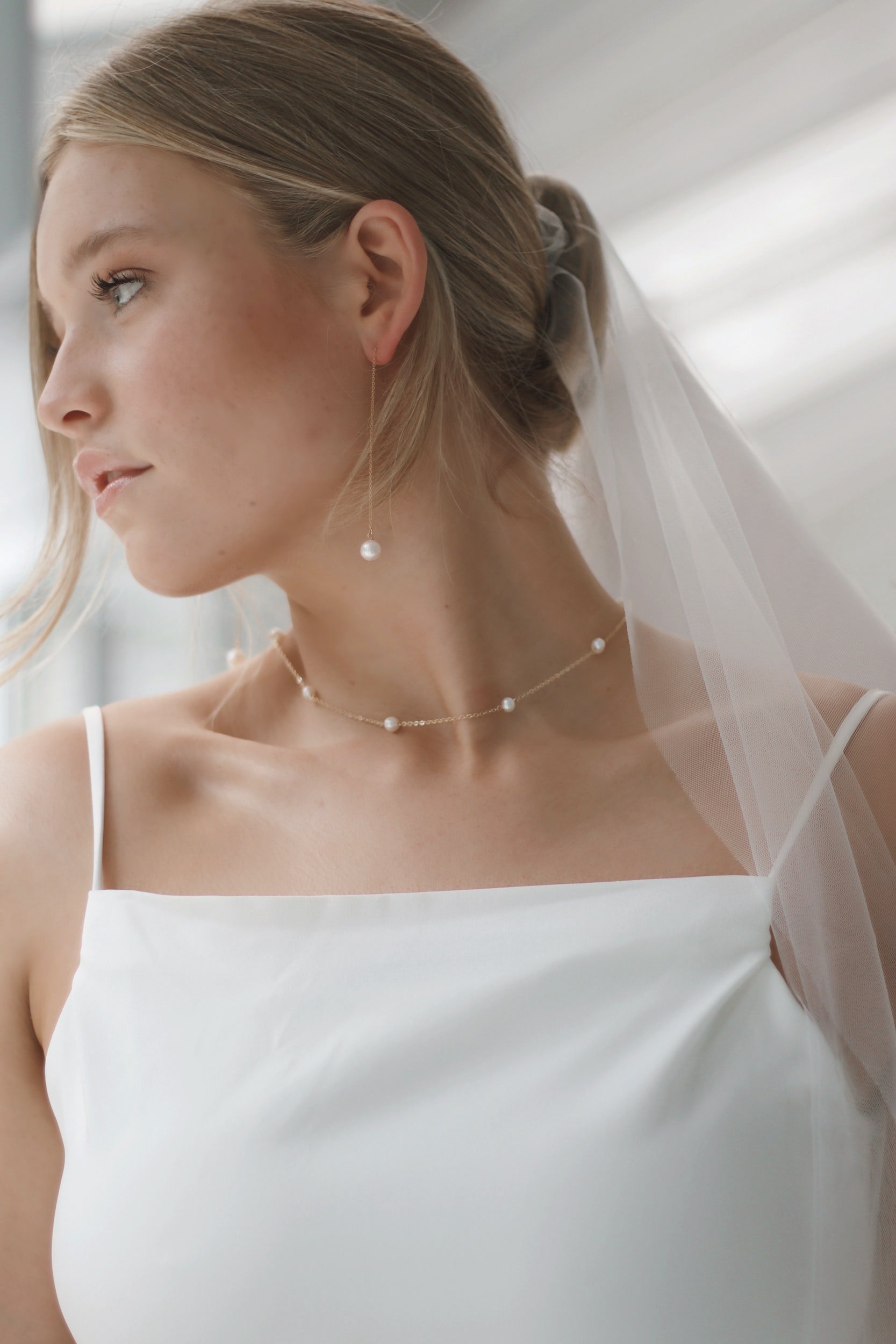 A young woman with blonde hair styled in a low updo, wearing a white dress with thin straps, a pearl necklace, and pearl drop earrings. She has a sheer veil attached to her hair and is looking to the left.