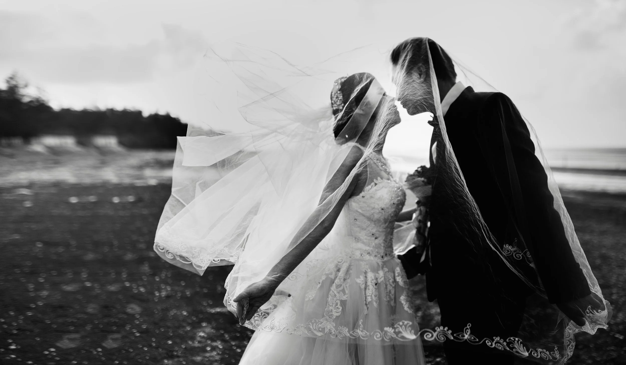 Black and white photo of a bride and groom with their foreheads touching, standing close on a beach during sunset, both wearing wedding attire and veils.