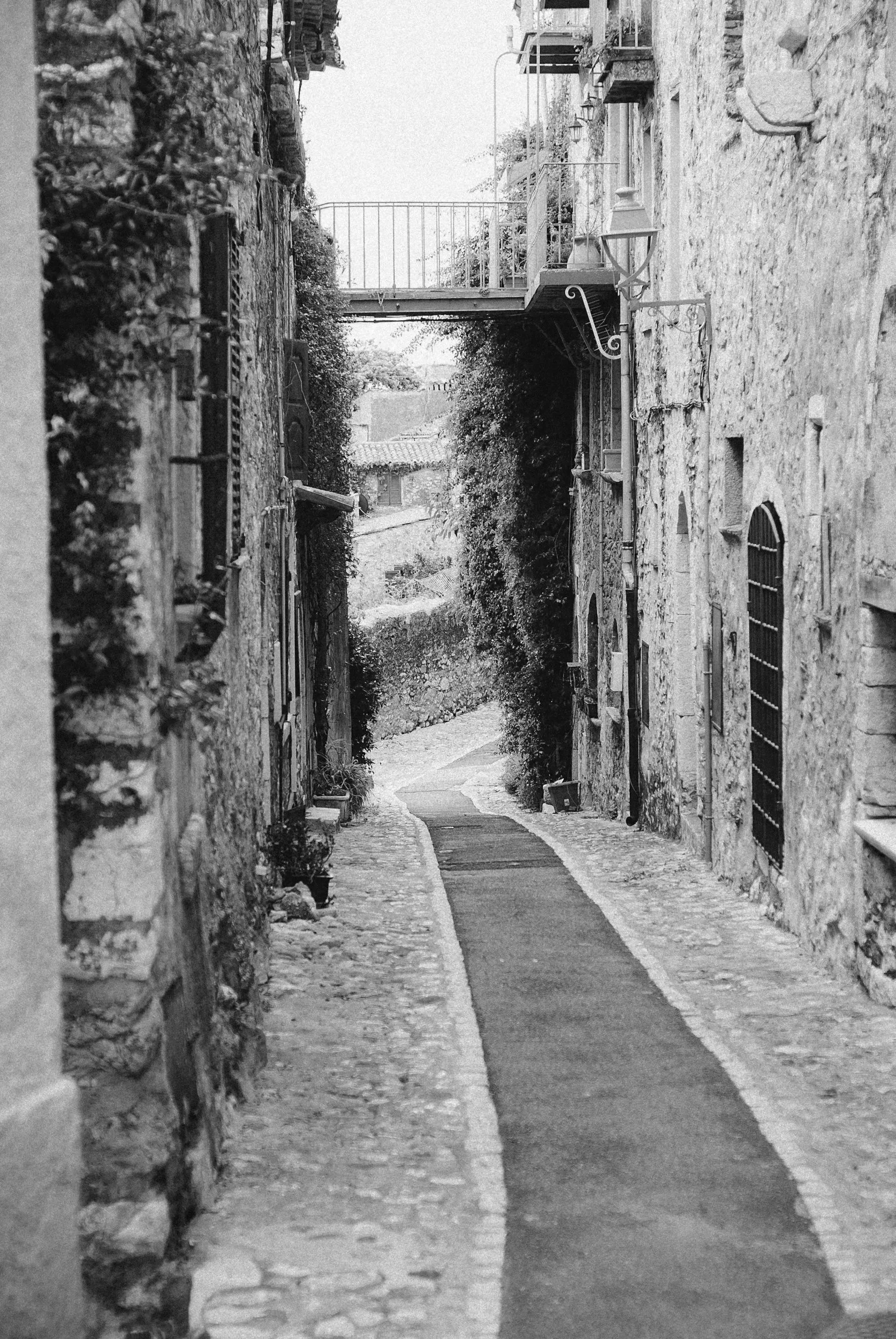 A narrow cobblestone alleyway between old stone buildings, with small plants and a railing on the upper level, in a rustic European village.