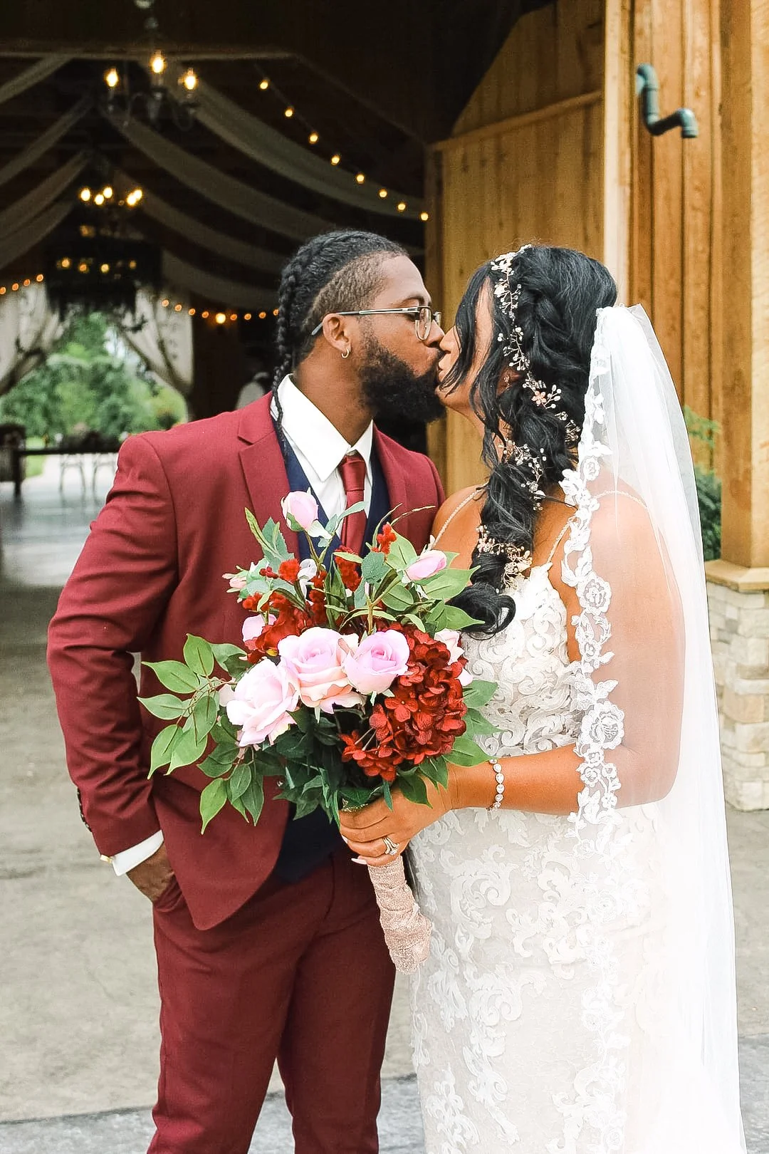 A couple is sharing a kiss at their wedding. The groom is wearing a red suit with a white shirt, red tie, and glasses, and has a beard and dreadlocks. The bride is dressed in a white lace wedding gown with floral detailing, holding a bouquet of pink and red flowers, with long black hair decorated with flowers and a veil.
