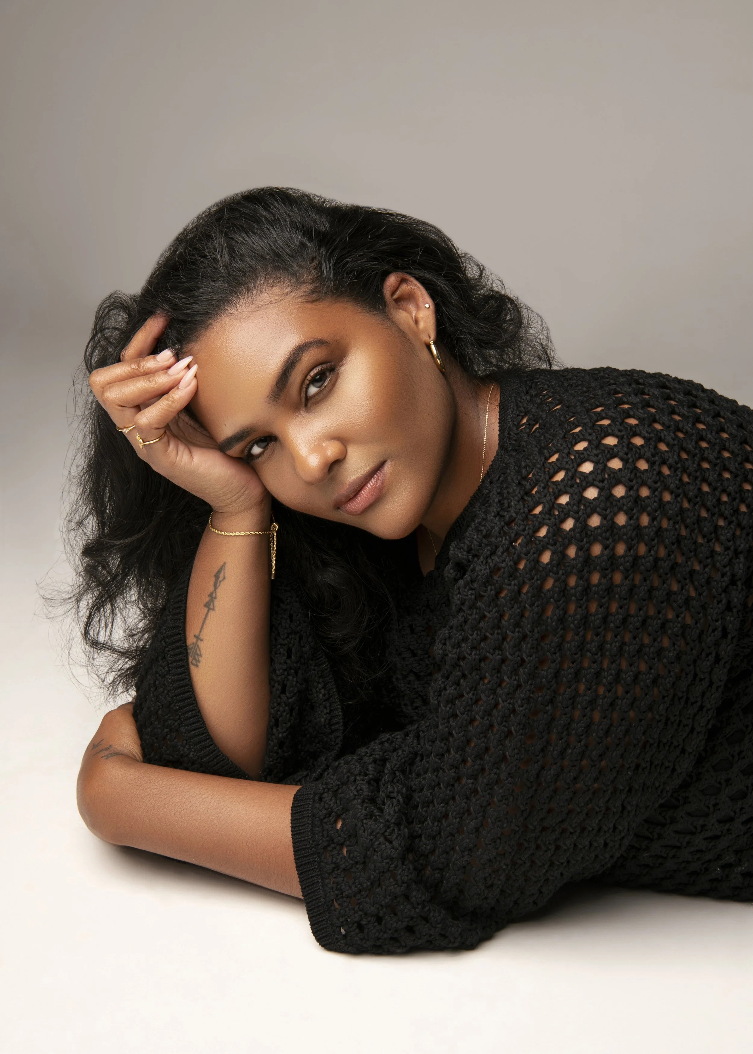 A woman with dark, curly hair resting her head on her hand. She is wearing gold hoop earrings, a gold necklace, and rings. She has a tattoo on her arm and is dressed in a black, open-knit sweater against a light gray background.