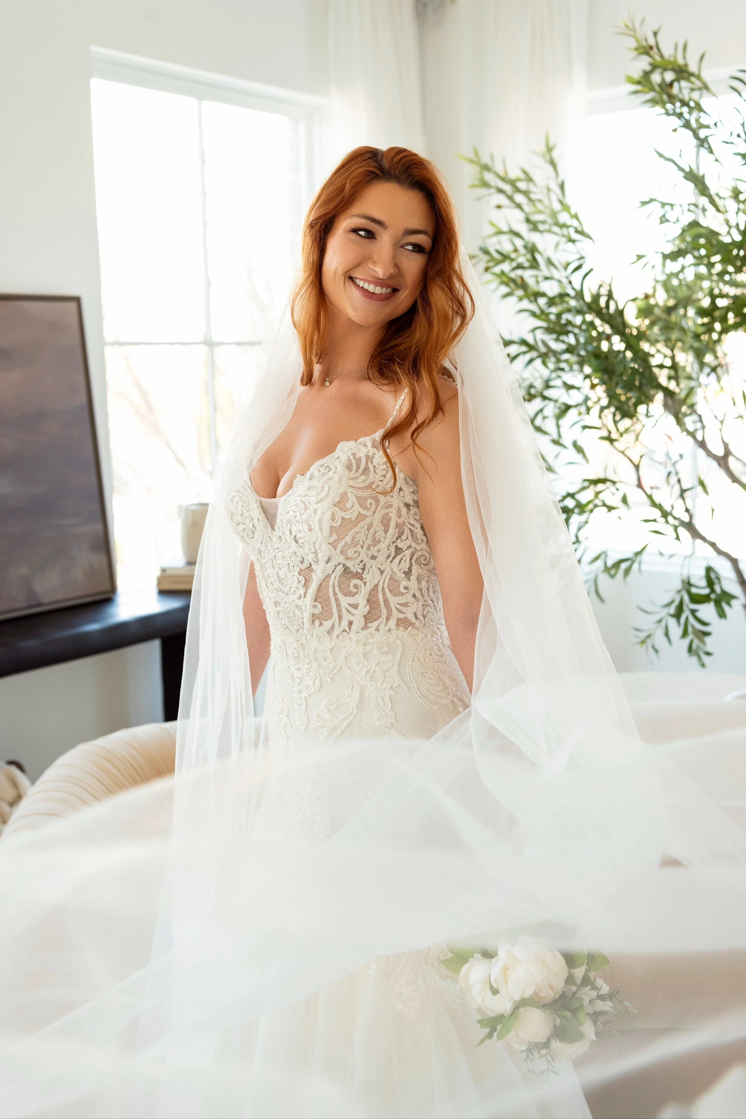 A smiling red-haired woman in a wedding dress with a veil standing inside a bright room with large windows and a plant in the background.