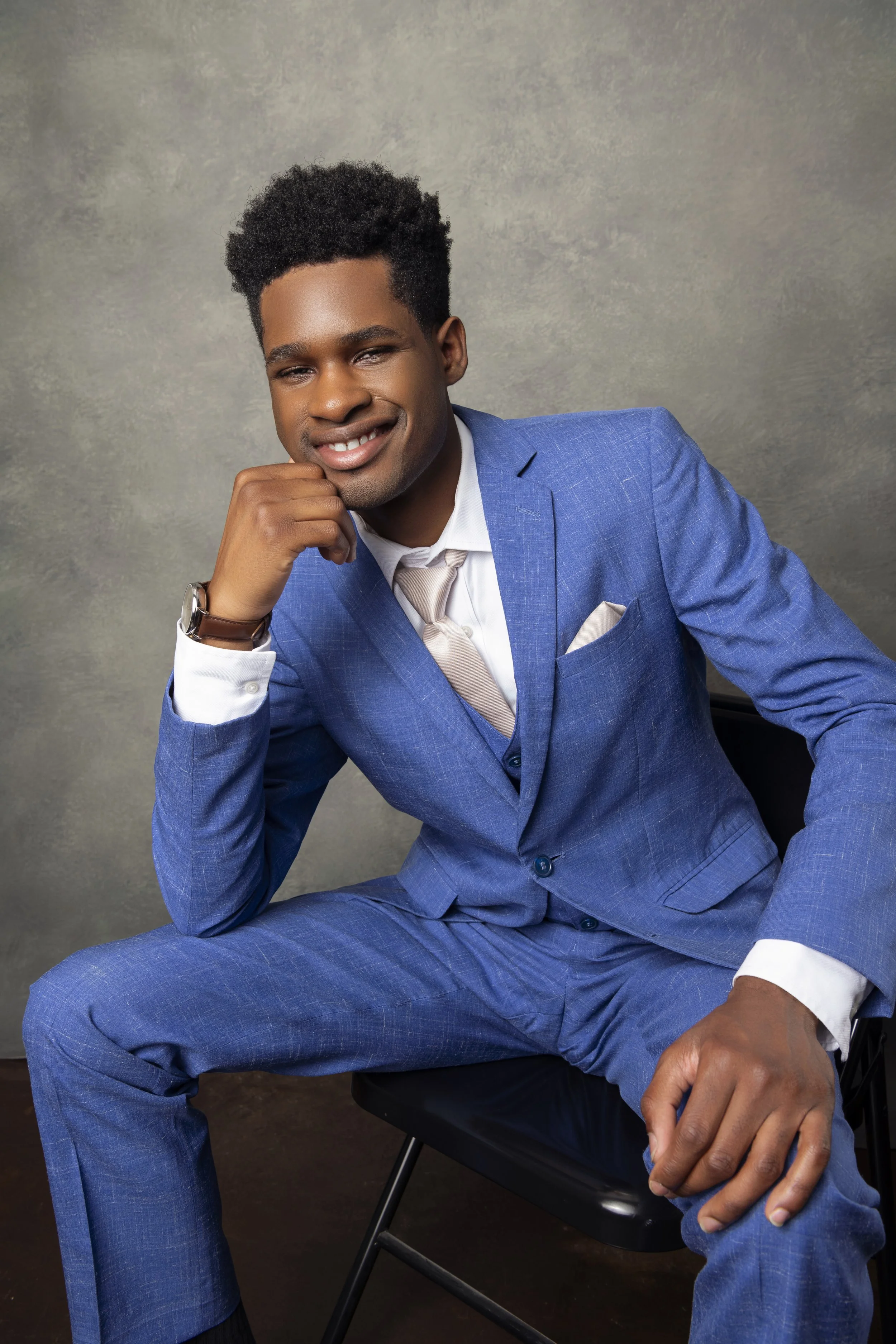 A young man with dark skin and curly hair is sitting on a black chair, wearing a blue suit, white shirt, and beige tie. He has a watch on his left wrist and is resting his chin on his left hand, smiling slightly. The background is a neutral gray text