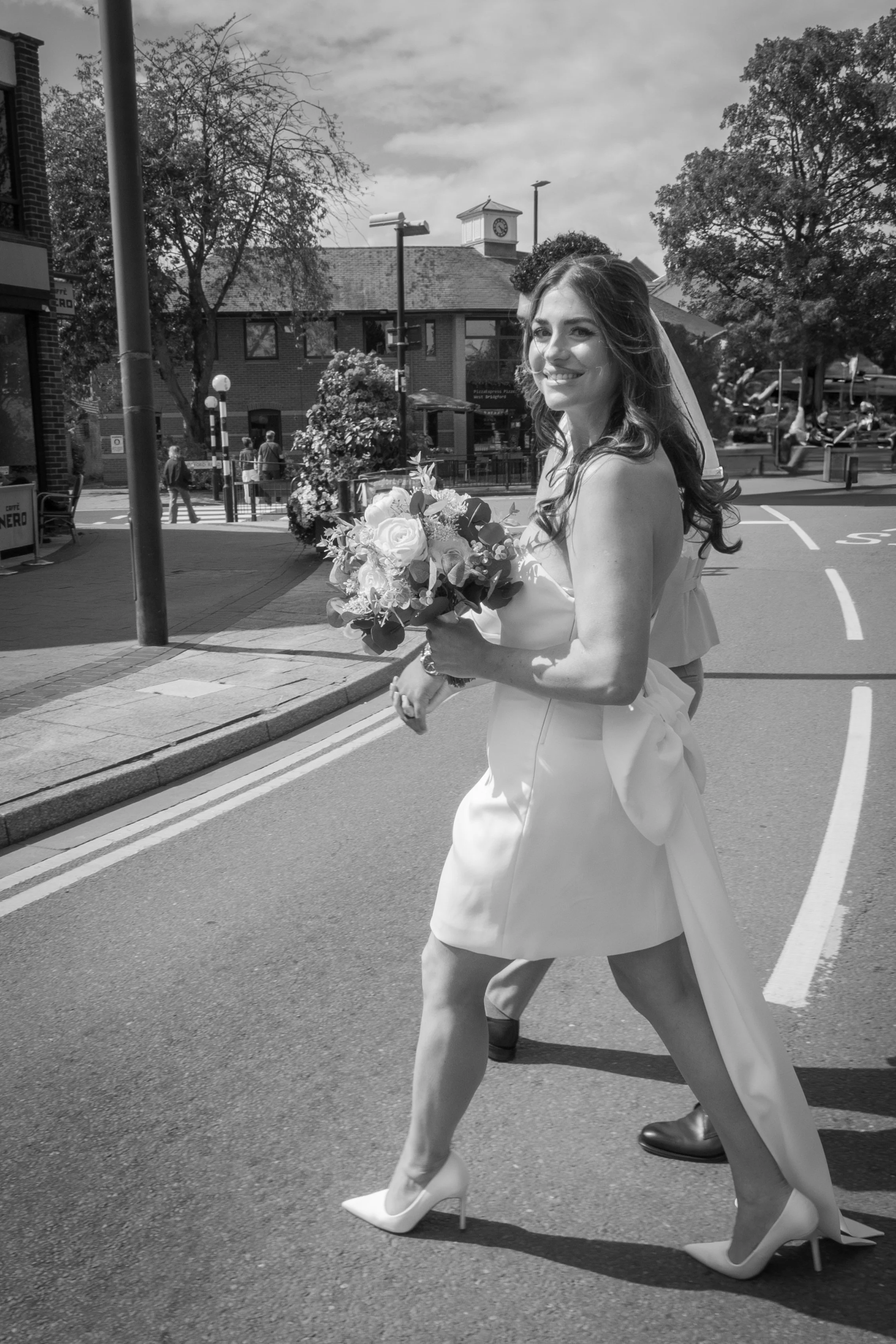 A smiling bride in a wedding dress holding a bouquet of flowers, walking on a city street.