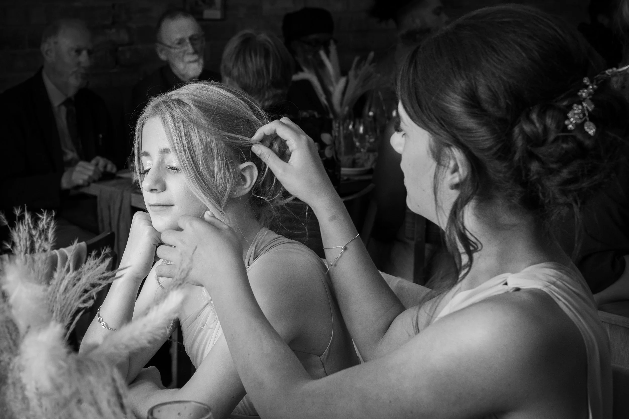 A woman decorates a young girl's hair at a social gathering, with people seated at tables in the background.
