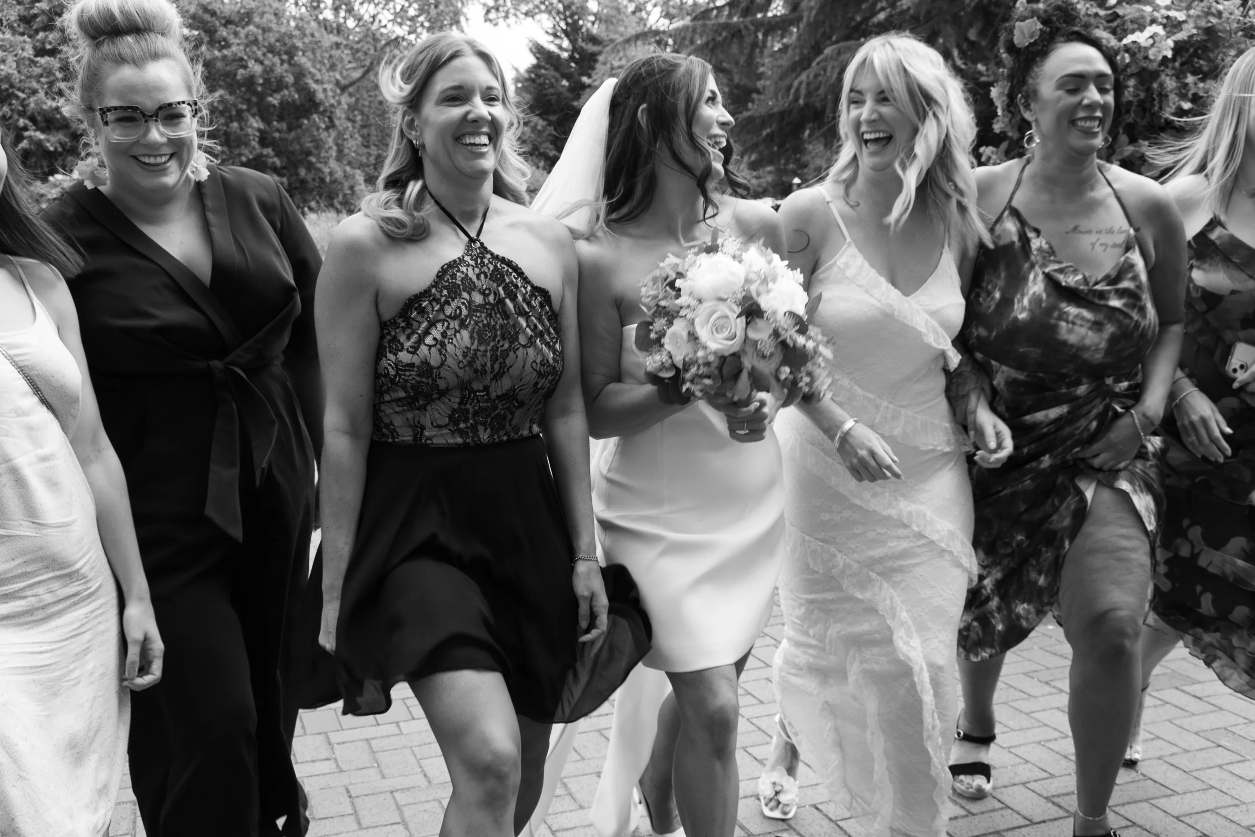 Black and white photo of a group of women walking outdoors, celebrating a wedding. The bride, wearing a white wedding dress and holding a bouquet, is smiling. Other women are dressed in various elegant outfits, also smiling and enjoying the moment.