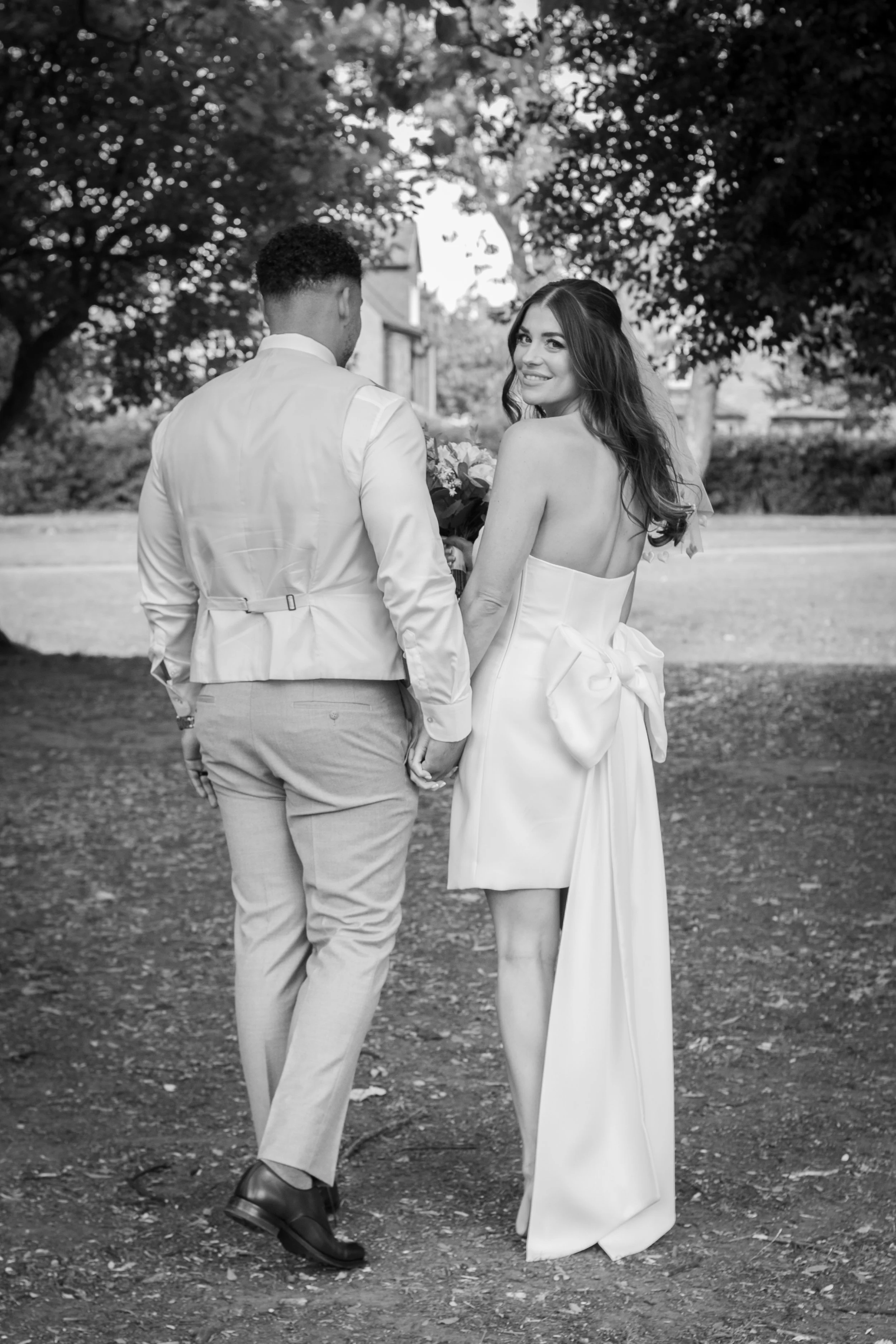 Black and white photo of a bride and groom holding hands, walking outdoors under trees. The bride is smiling and looking over her shoulder, wearing a strapless dress with a bow. The groom, seen from the back, is wearing a light-colored vest and pants