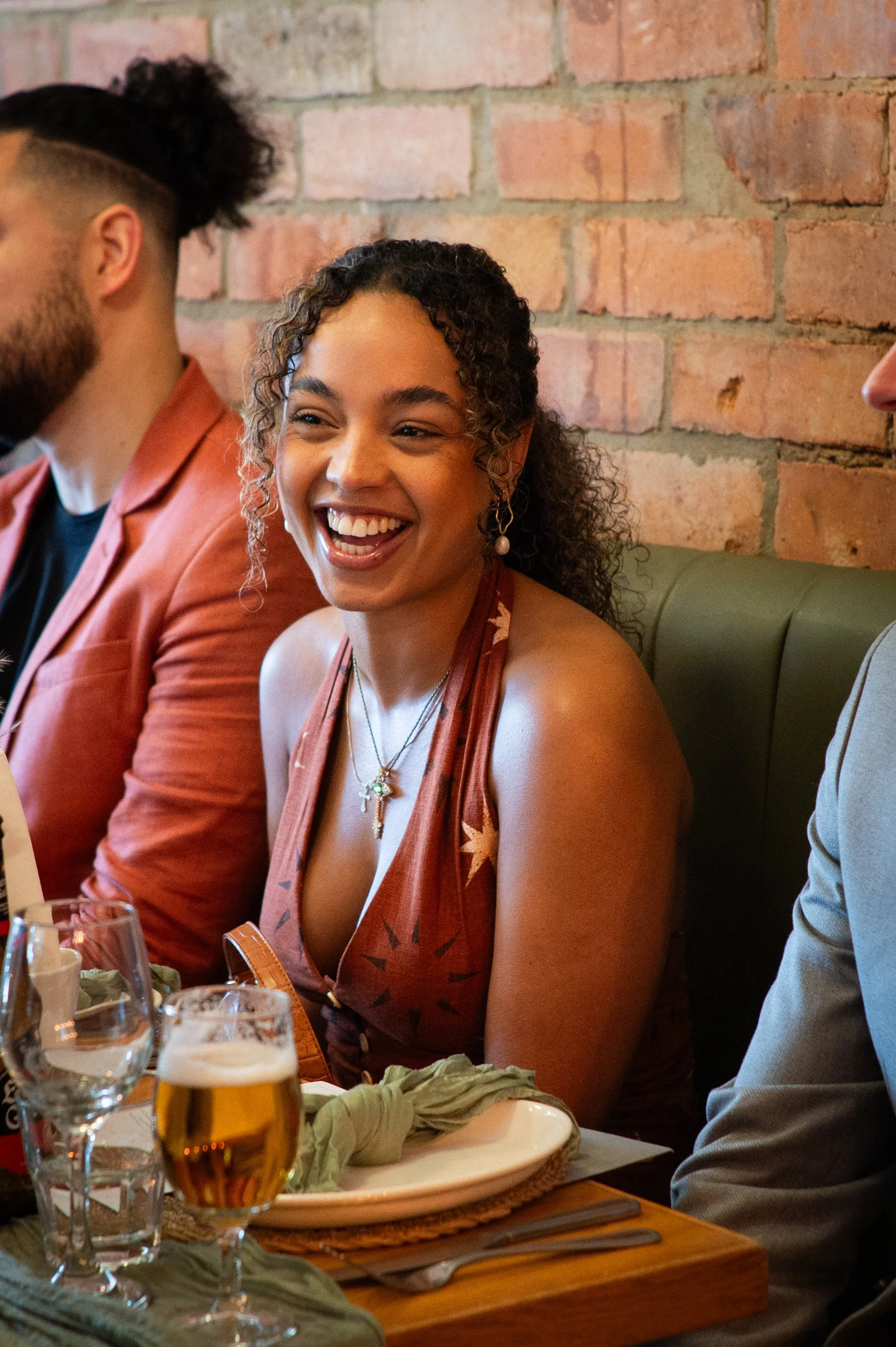 A woman with curly hair smiling while sitting at a restaurant table with other people, with a brick wall in the background.
