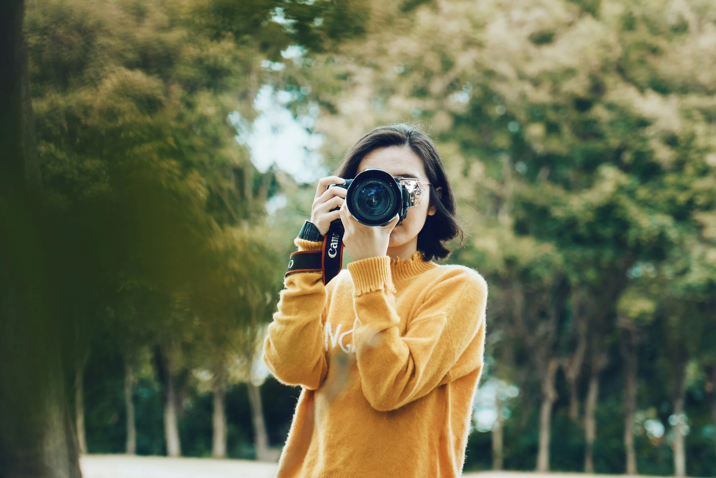 A young woman with dark hair wearing an orange sweater, holding a camera up to her face, taking a photo outdoors in a park with green trees in the background.