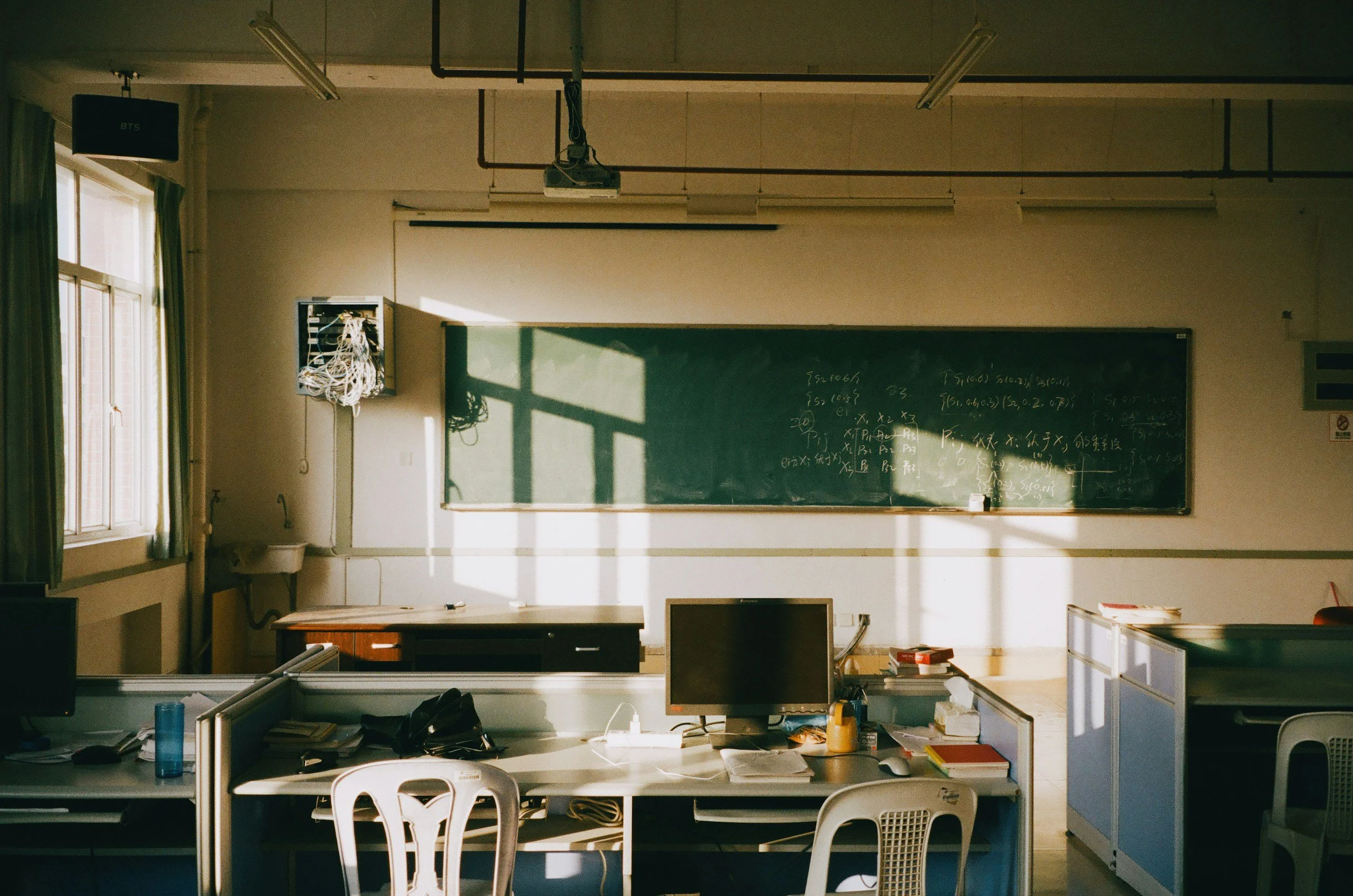 Empty classroom with sunlight streaming through windows onto desks, computer, and chalkboard with chalk writing.